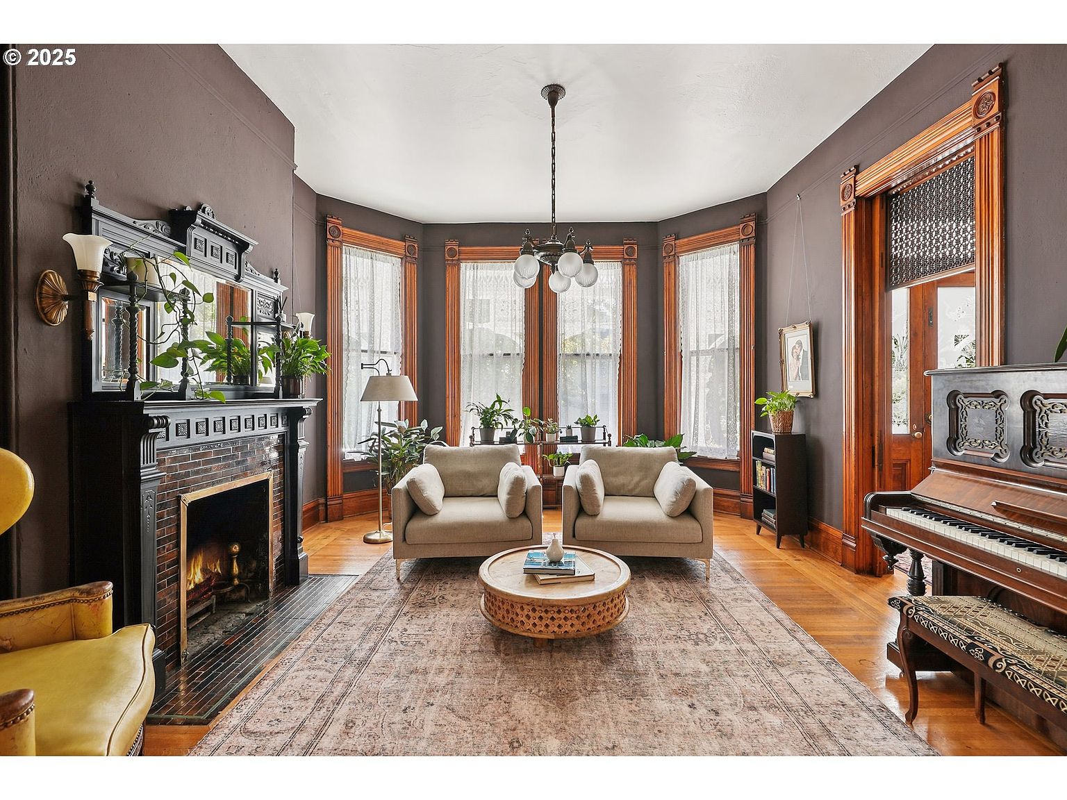 This is a beautifully appointed living room featuring dark walls, ornate wood trim, and hardwood floors covered by a large area rug. The room is furnished with two beige sofas, a round coffee table, and a grand piano, creating a warm and inviting atmosphere. A fireplace with an elaborate mantel adds a focal point, while large windows provide ample natural light.