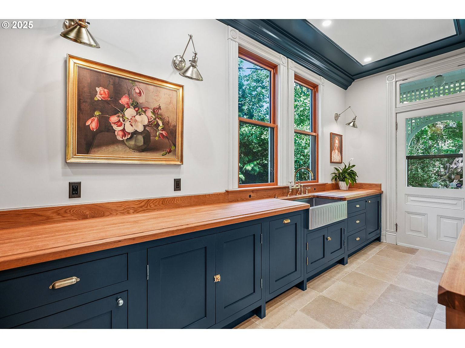 This interior shot showcases a charming kitchen with dark blue cabinetry and a warm wood countertop. A farmhouse sink sits beneath two windows, providing natural light. The room is decorated with a framed painting and sconce lighting, adding to the classic and elegant feel of the space.