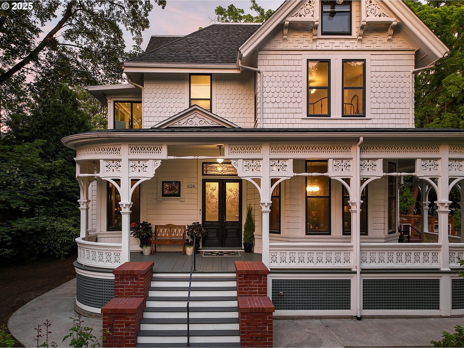 This is a charming front view of a two-story Victorian-style house. The house features a large, ornate porch with decorative columns and railings, leading to a grand entrance with double doors. The exterior is painted in a light color, and the roof is dark, creating a classic and inviting curb appeal.