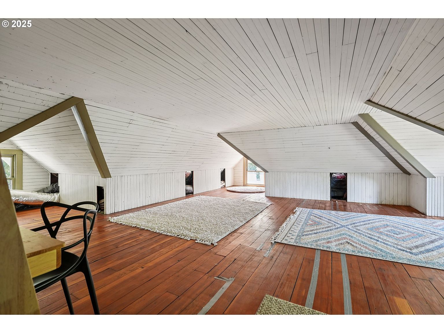 This is an interior shot of an attic space featuring a white painted plank ceiling and walls, with exposed beams. The floor is made of stained wood planks, and several area rugs are scattered throughout the room. The space is illuminated by natural light coming through small windows, creating a cozy and inviting atmosphere.