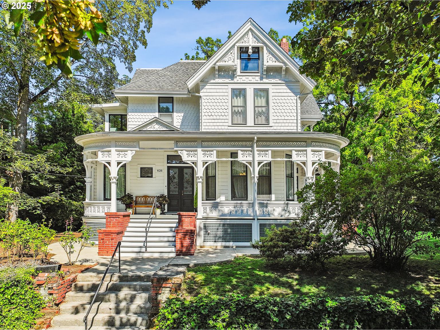 This is a charming front view of a two-story Victorian-style house with white siding and intricate architectural details. The house features a curved porch with decorative columns, a prominent gable with a window, and a well-maintained front yard with mature trees and landscaping. The inviting entryway is accessed by a set of stairs with brick accents, creating a welcoming and picturesque scene.