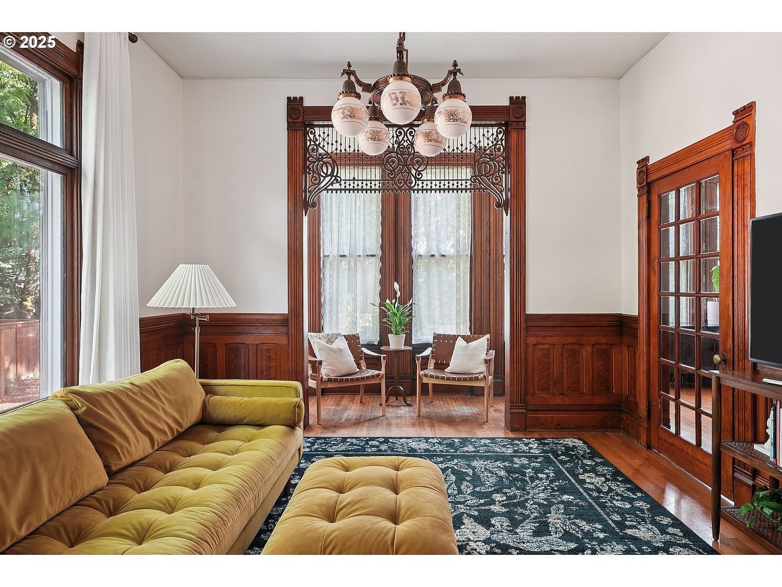 This is an interior shot of a living room featuring a vintage aesthetic. The room is furnished with a mustard-colored sofa and ottoman, a dark blue patterned rug, and two wooden chairs with cushions. The room has dark wood trim and paneling, a decorative window frame, and a unique chandelier, creating a warm and inviting atmosphere.