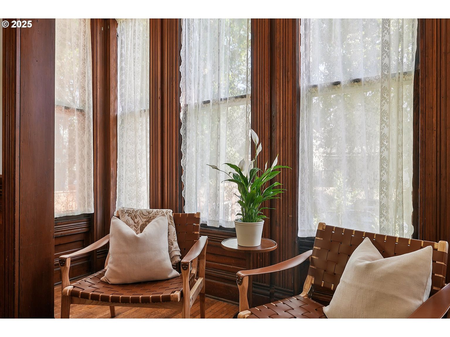 This sunroom features rich, dark wood paneling and large windows covered with sheer, lacy curtains, creating a bright and airy atmosphere. Two woven leather chairs with plush pillows invite relaxation, while a small side table holds a potted peace lily, adding a touch of nature. The room exudes a sense of classic elegance and tranquility.