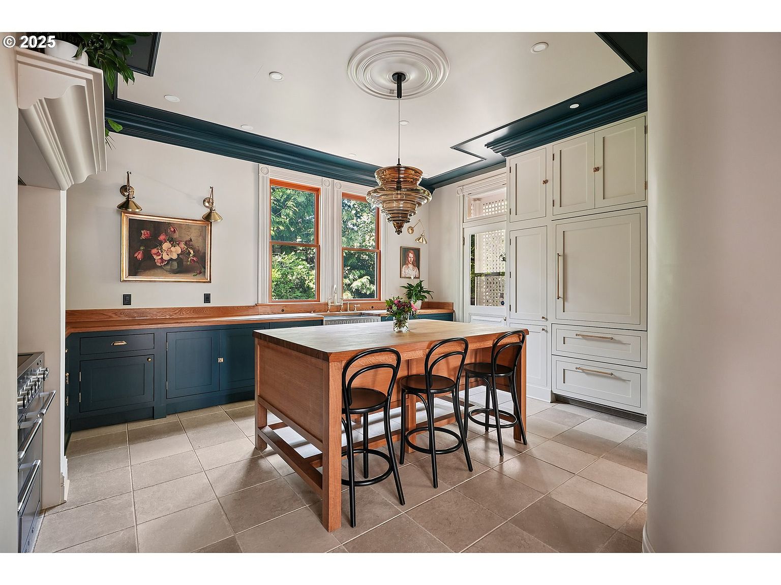 This is a well-lit kitchen featuring a central wooden island with bar stools, dark blue lower cabinets, and white upper cabinets that extend to the ceiling. The kitchen also has a decorative ceiling medallion with a modern chandelier, and a large window provides natural light. The overall impression is a blend of classic and contemporary design.