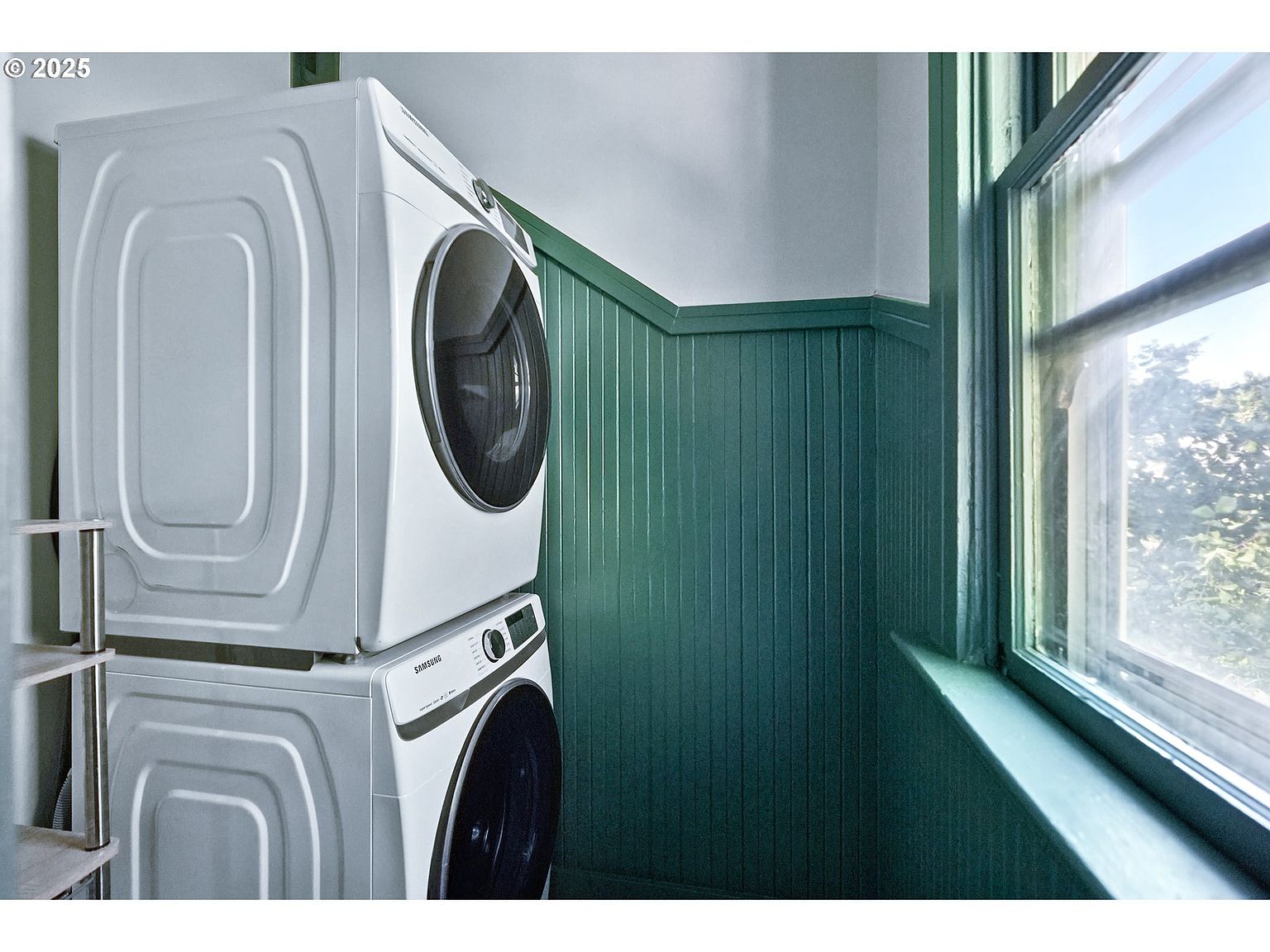 This laundry room features a stacked Samsung washer and dryer set, maximizing space. The room is decorated with green wainscoting and trim, complemented by white walls above. A window provides natural light, creating a clean and functional space.