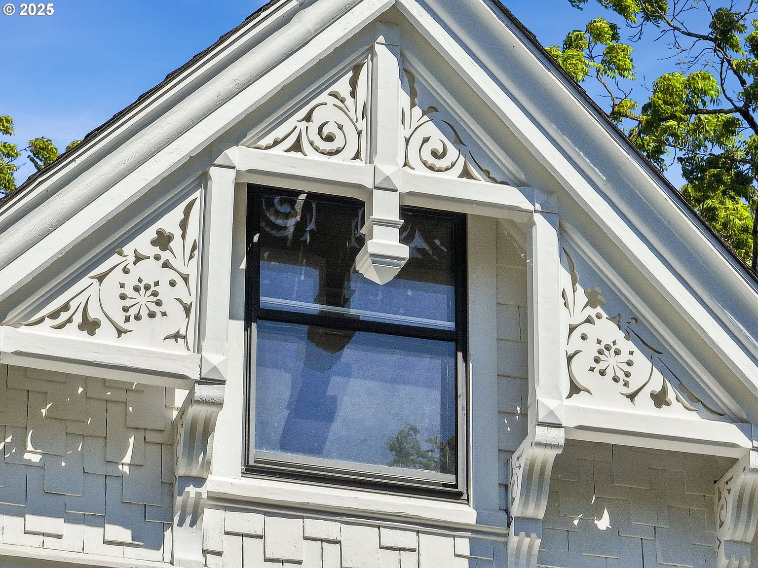 This image showcases a close-up of an architectural detail, specifically a gable window with ornate trim. The window is framed by decorative woodwork featuring floral and scroll patterns, adding a touch of elegance and craftsmanship to the exterior. The white color scheme enhances the intricate details and creates a visually appealing focal point.
