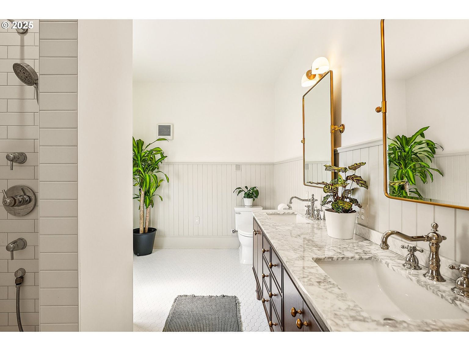 This is a well-lit primary bathroom featuring a double vanity with a marble countertop and dark wood cabinetry. Two gold-framed mirrors hang above the sinks, complemented by sconce lighting. The bathroom also includes a shower with subway tile and a separate toilet area, with plants adding a touch of nature to the space.