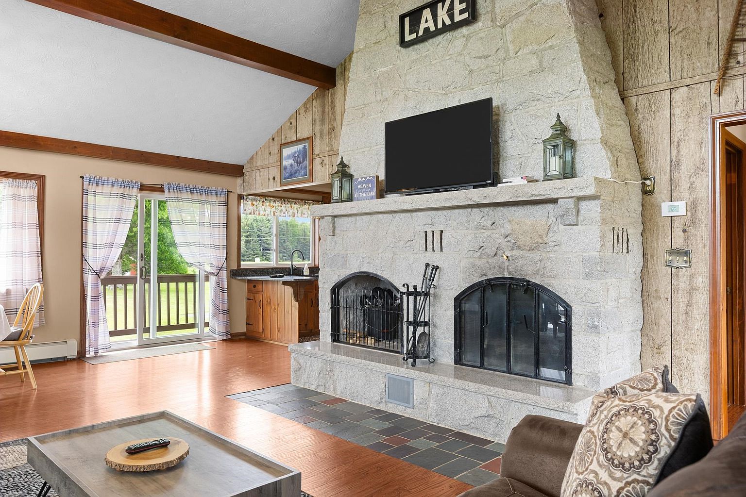 This cozy living room features a prominent stone-clad chimney breast with dual arched fireplace openings and a central television mount. The space is accented by rustic wood-paneled walls, a vaulted ceiling with exposed beams, and a sliding glass door that opens to an outdoor deck. A neutral-toned sofa with patterned pillows sits in the foreground, creating a warm and inviting atmosphere.
