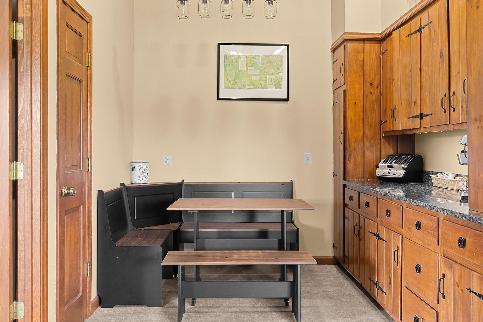 This cozy breakfast nook features a built-in corner bench with a matching table and bench seating, creating an intimate dining area. To the right, a wall of warm-toned wood cabinetry provides ample storage and counter space, complemented by a dark stone countertop and a toaster. The space is illuminated by a row of five glass pendant lights hanging from the ceiling, and a framed map hangs on the wall above the seating area.