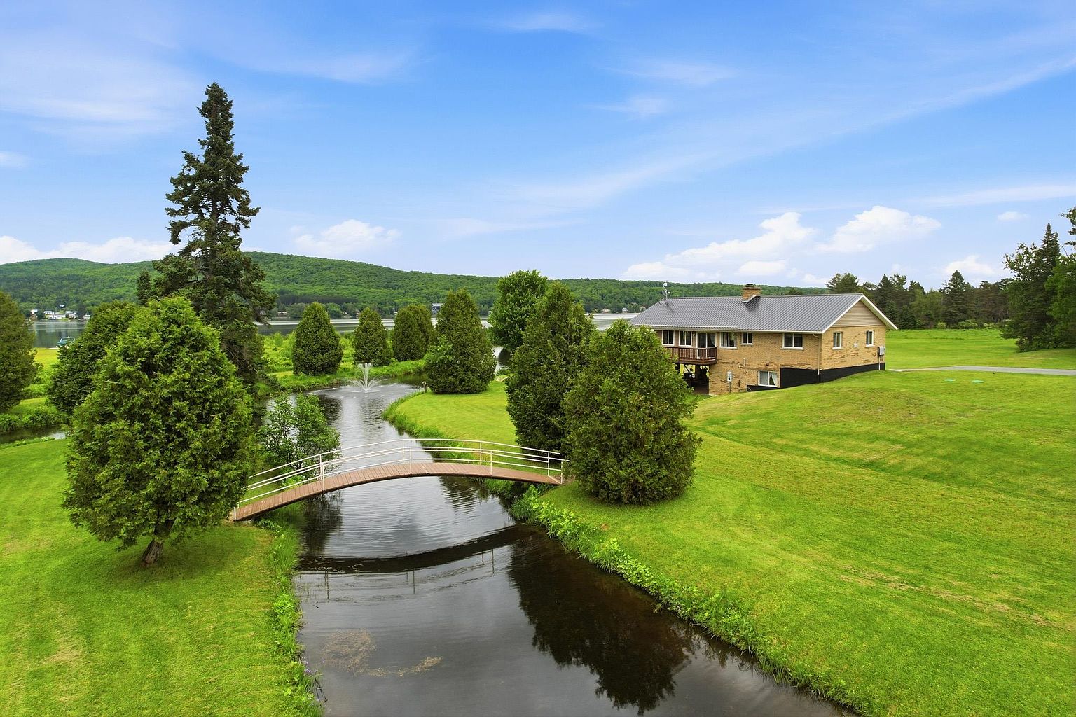 This elevated aerial perspective captures a sprawling property featuring a brick-clad residence situated on a lush, expansive lawn. A charming arched footbridge spans a tranquil stream that flows through the foreground, complete with a small water fountain. The scene is framed by mature evergreen trees and rolling hills in the distance, creating a serene and picturesque rural setting.