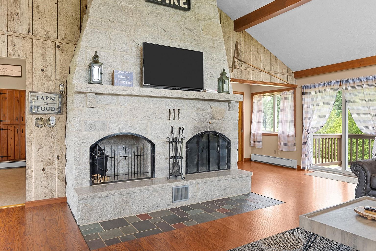 This living room features a prominent stone-clad chimney breast that serves as the focal point, housing a television and two arched fireboxes. The space is accented by rustic wood-paneled walls, a vaulted ceiling with exposed beams, and a slate-tiled hearth. A sliding glass door leads to an outdoor deck, allowing natural light to fill the room, which is furnished with a neutral-toned sofa and a textured area rug.