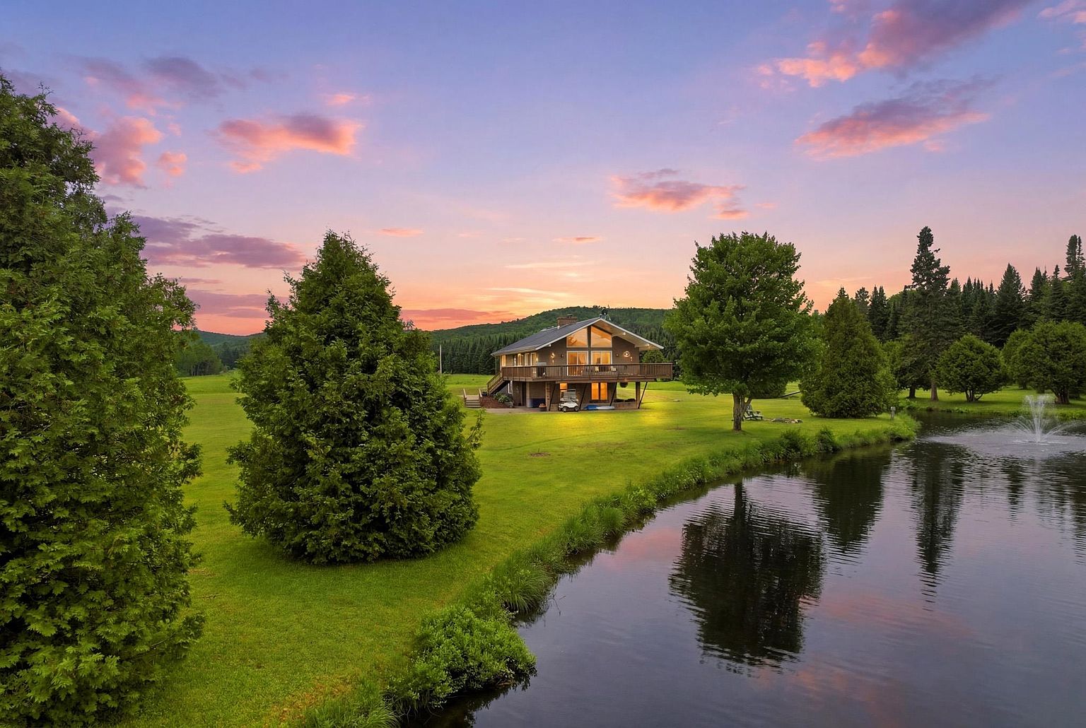This elevated, wide-angle shot captures a charming two-story home nestled in a lush, expansive green landscape at sunset. The house features a prominent deck overlooking a serene pond with a fountain, while the surrounding property is dotted with mature trees and rolling hills in the distance. The warm, golden light of the setting sun creates a peaceful and inviting atmosphere, highlighting the home's architectural integration with its natural surroundings.