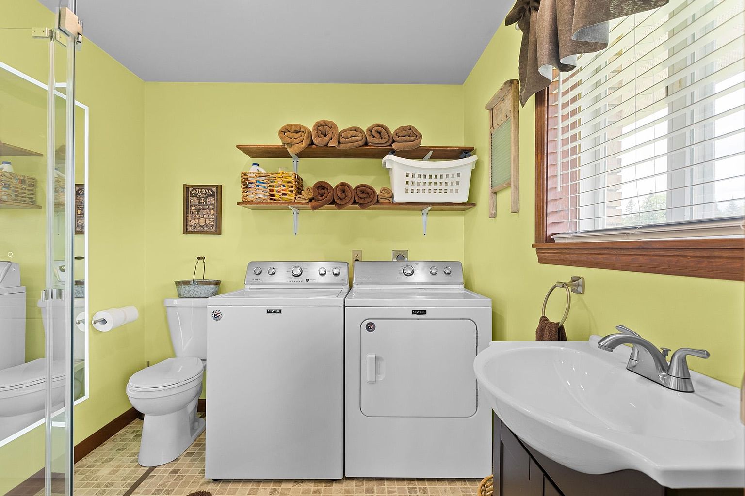 This multi-functional laundry room features bright yellow walls and a tiled floor, housing a side-by-side Maytag washer and dryer set. Above the appliances, two wooden shelves provide storage for rolled towels and a laundry basket, while a pedestal sink and a toilet are positioned to the side. A window with blinds allows natural light into the space, which also includes a decorative sign and a vintage-style washboard mounted on the wall.