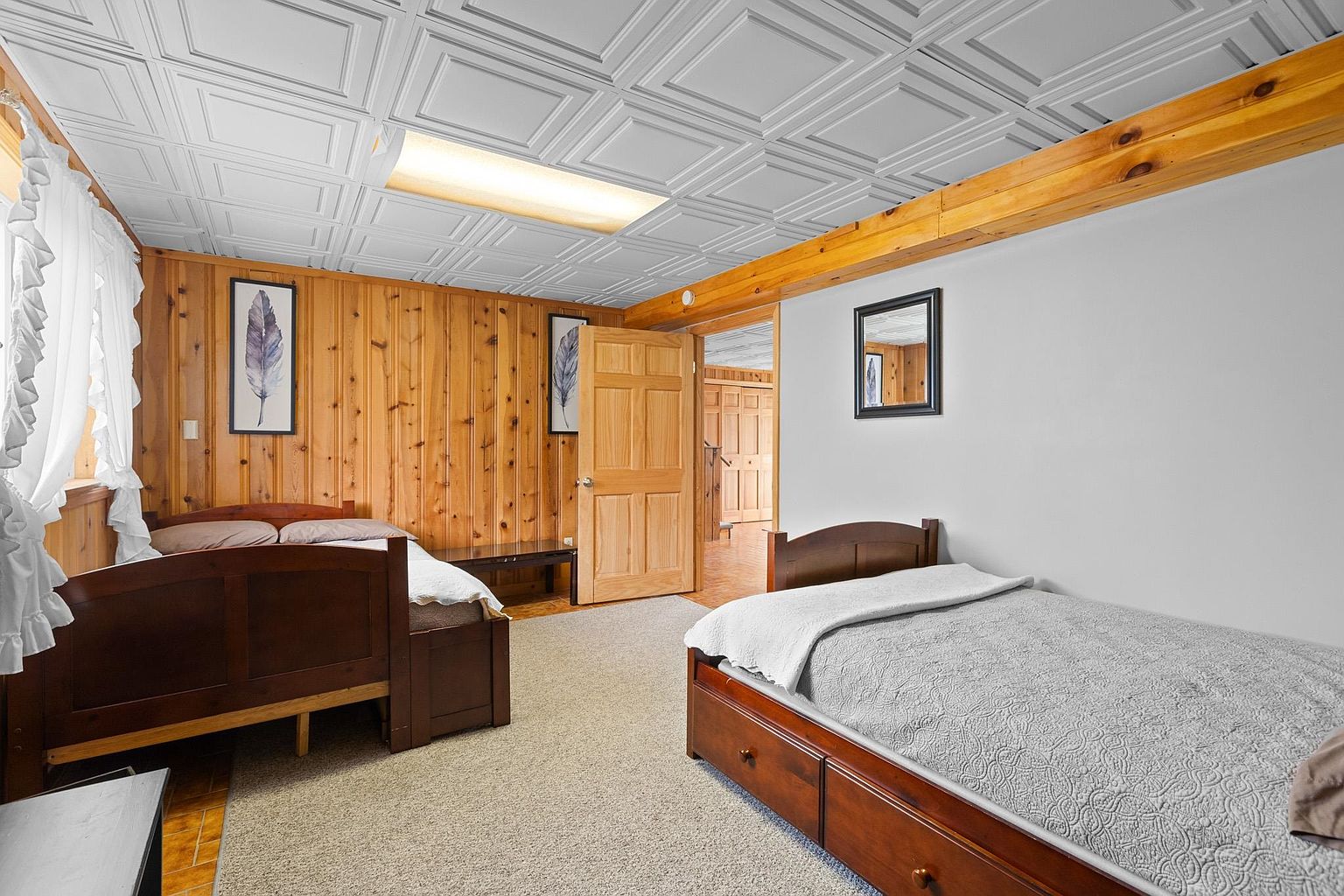 This bedroom features a rustic aesthetic with wood-paneled walls and a textured white ceiling. The room is furnished with two wooden bed frames, one positioned against the wood wall and the other against a neutral-toned wall, both dressed in simple bedding. A doorway leads into an adjacent room, and the space is illuminated by a central light fixture, creating a cozy and functional sleeping area.