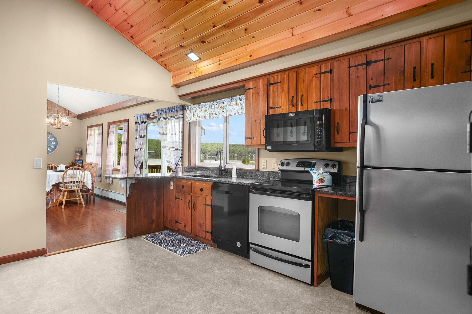 This kitchen features warm-toned wood cabinetry with rustic hardware, paired with dark stone countertops and stainless steel appliances. A vaulted wood-paneled ceiling adds architectural interest, while a window above the sink provides a view of the outdoors. The space flows openly into an adjacent dining area, creating a cohesive and inviting atmosphere.
