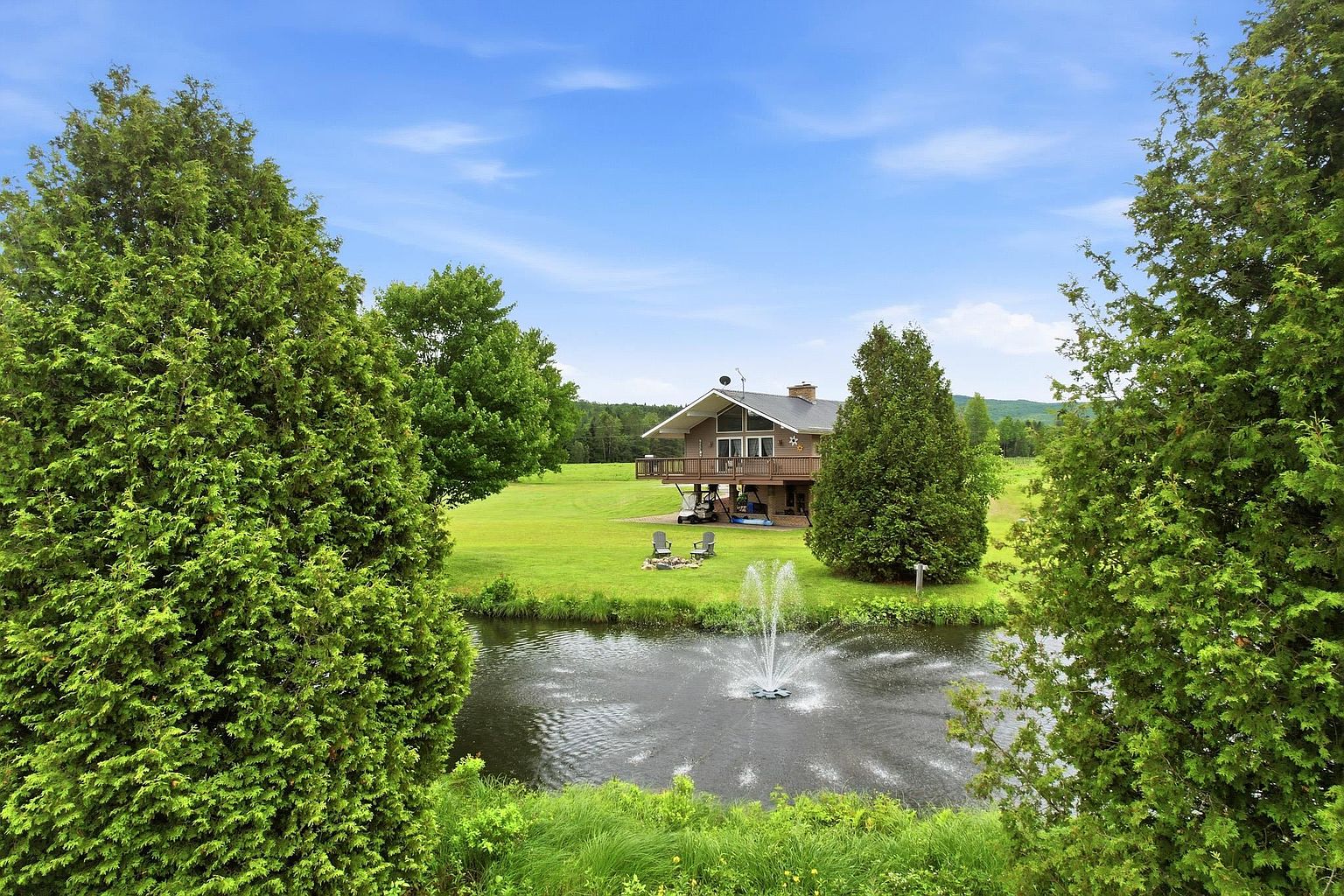 This scenic exterior view captures a multi-level home nestled in a lush, green landscape, framed by two large, mature evergreen trees in the foreground. A decorative water fountain sprays in a small pond, leading the eye toward the house, which features a prominent elevated deck and a walk-out lower level. The property is surrounded by expansive lawns and rolling hills, creating a serene and private atmosphere.
