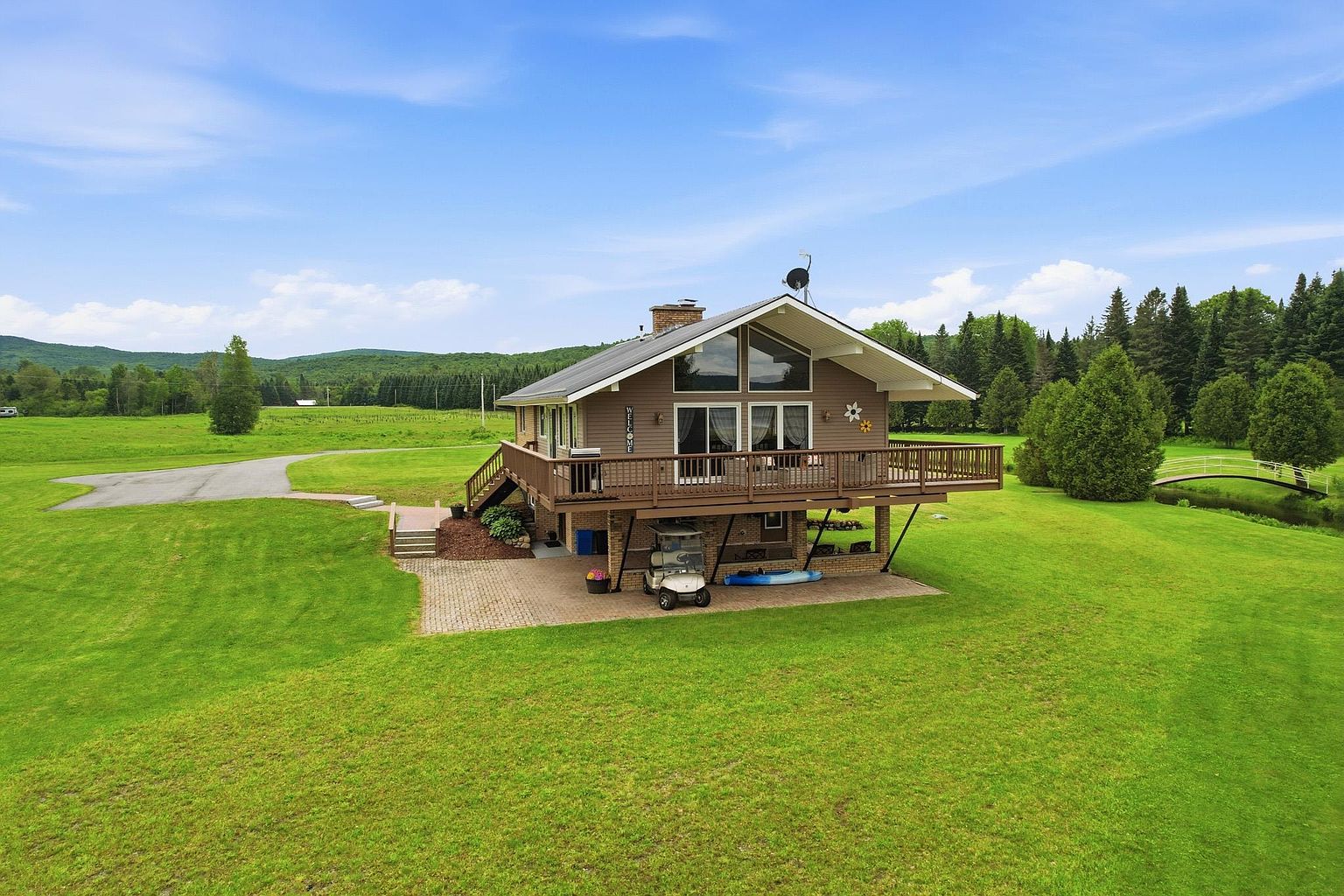 This elevated perspective captures a multi-level home set within a sprawling, manicured lawn. The house features a large wooden deck extending from the main living level, with a covered patio area underneath that houses a golf cart and recreational equipment. A small arched bridge crosses a stream in the background, emphasizing the expansive, park-like grounds surrounding the property.