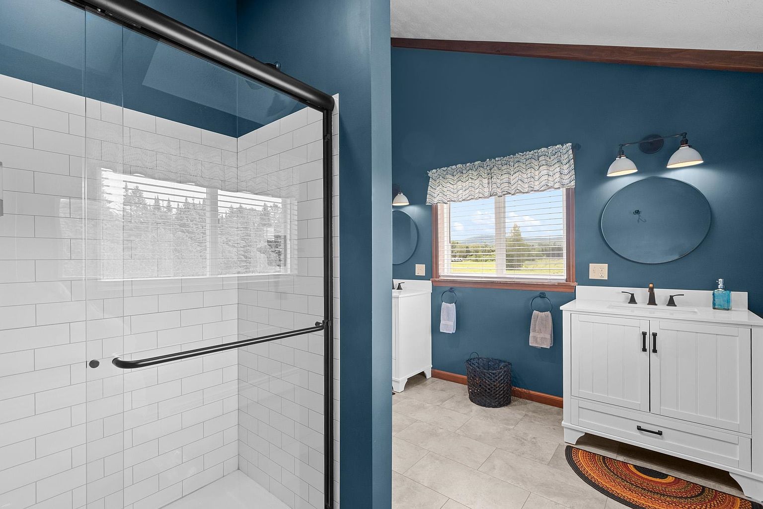 This bright bathroom features a walk-in shower with white subway tile and a black-framed glass enclosure. The space is painted in a deep blue, which contrasts with the white vanity and light-colored tile flooring. A window with a patterned valance provides natural light, while dual round mirrors and wall-mounted light fixtures complete the vanity area.