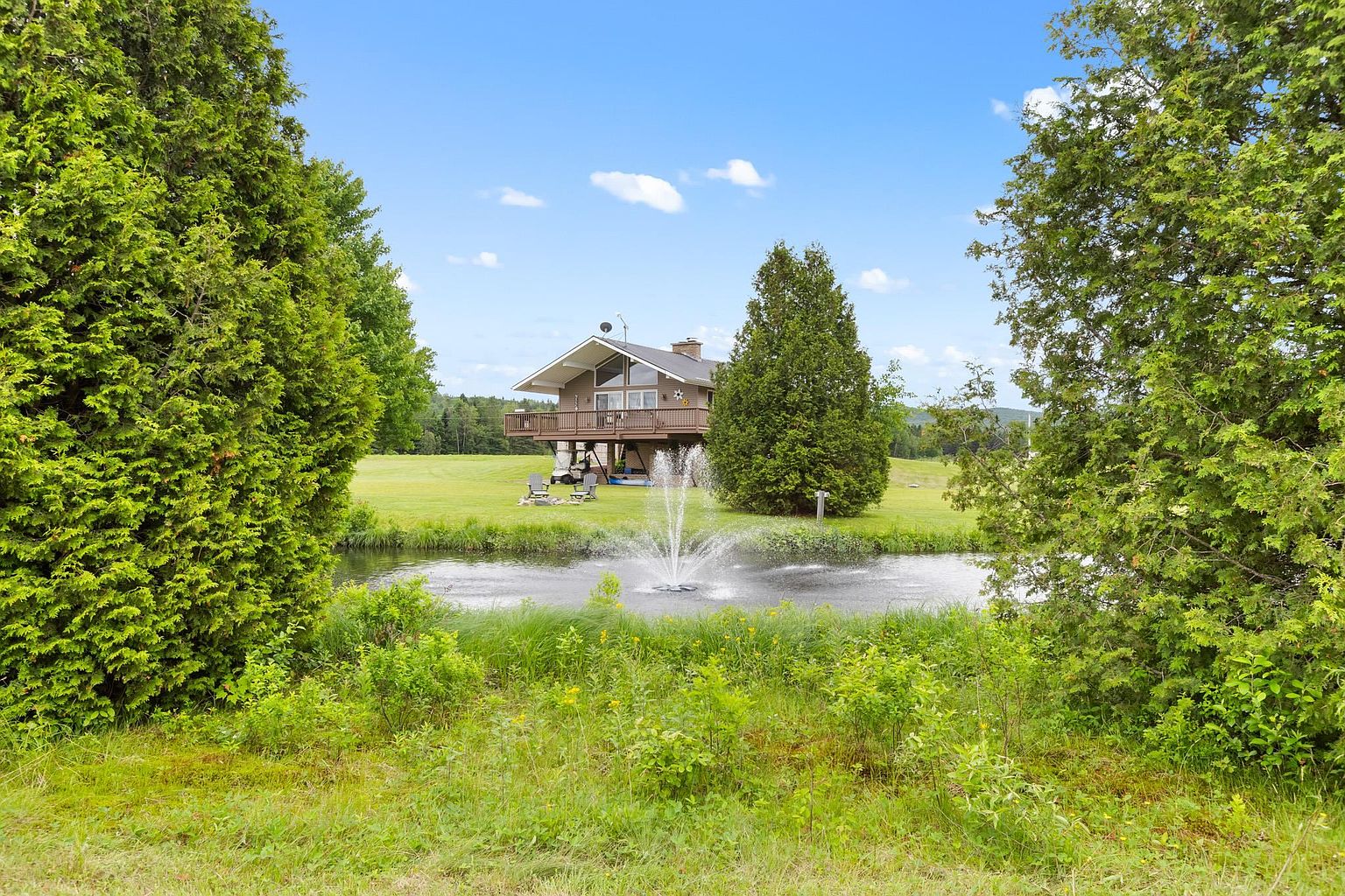 This rear view of a multi-level home showcases a spacious wooden deck overlooking a manicured lawn and a tranquil pond with a central fountain. The property is framed by lush, mature evergreen trees, creating a private and serene atmosphere. The house features a classic architectural style with large windows that capture the natural light and scenic surroundings.