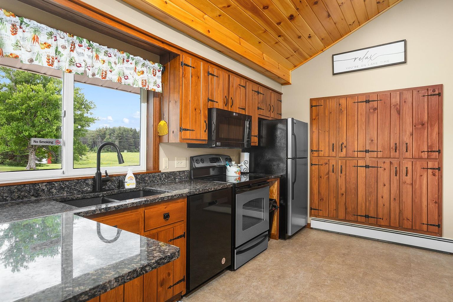 This rustic-style kitchen features warm wood cabinetry, dark stone countertops, and a black appliance suite. A large window above the sink provides a bright view of the outdoors, while a wooden plank ceiling adds a cozy, cabin-like aesthetic. The space is completed by a large, wood-paneled pantry or storage wall and a decorative sign, creating a welcoming and functional atmosphere.