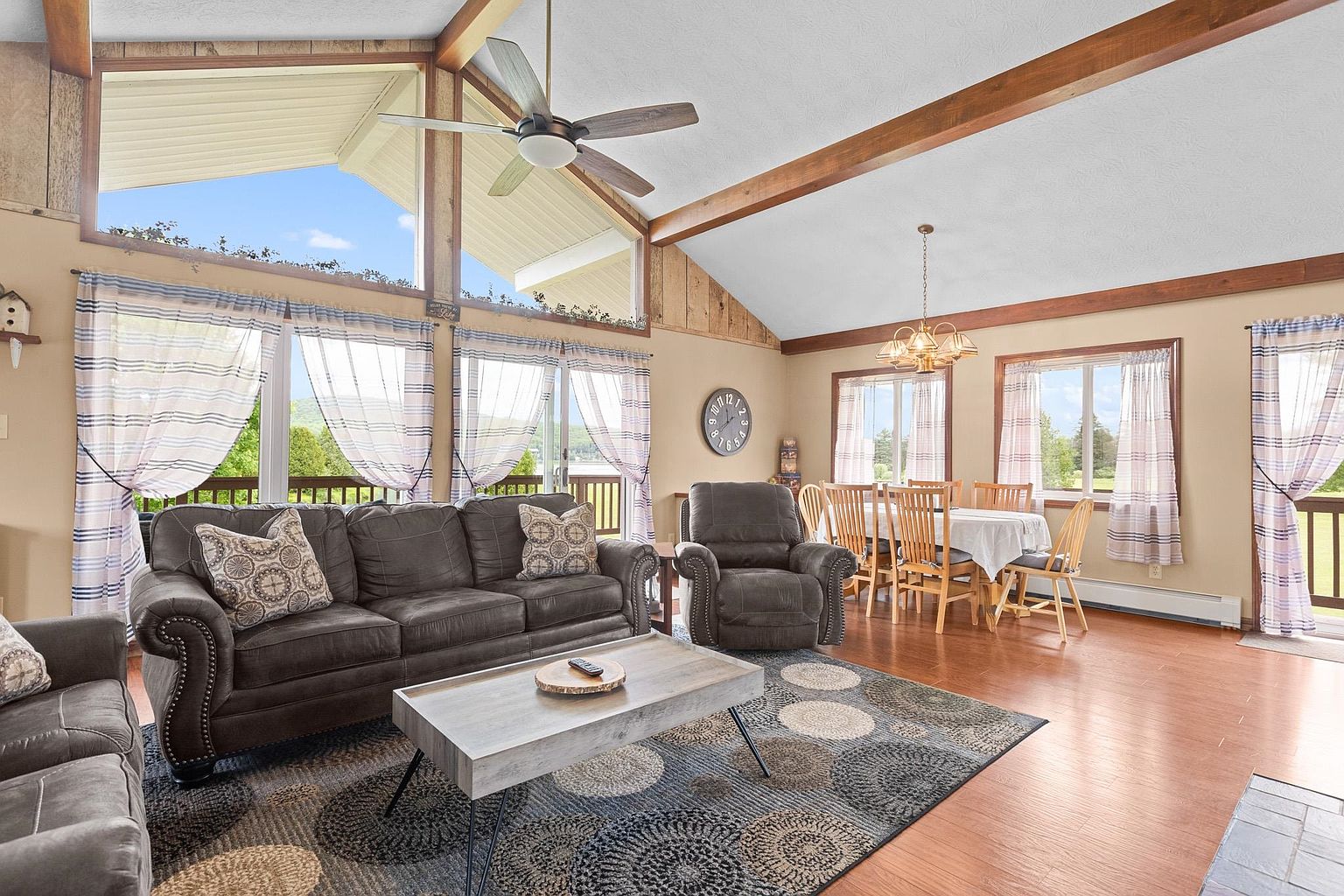This open-concept living and dining area features vaulted ceilings with exposed wooden beams and a large wall of windows that provide ample natural light. The living space is anchored by a dark sofa and matching recliner set atop a circular-patterned area rug, while the adjacent dining area includes a wooden table and chairs under a traditional chandelier. The room has a warm, inviting atmosphere with light-toned walls and wood-look flooring, creating a cohesive and comfortable living environment.