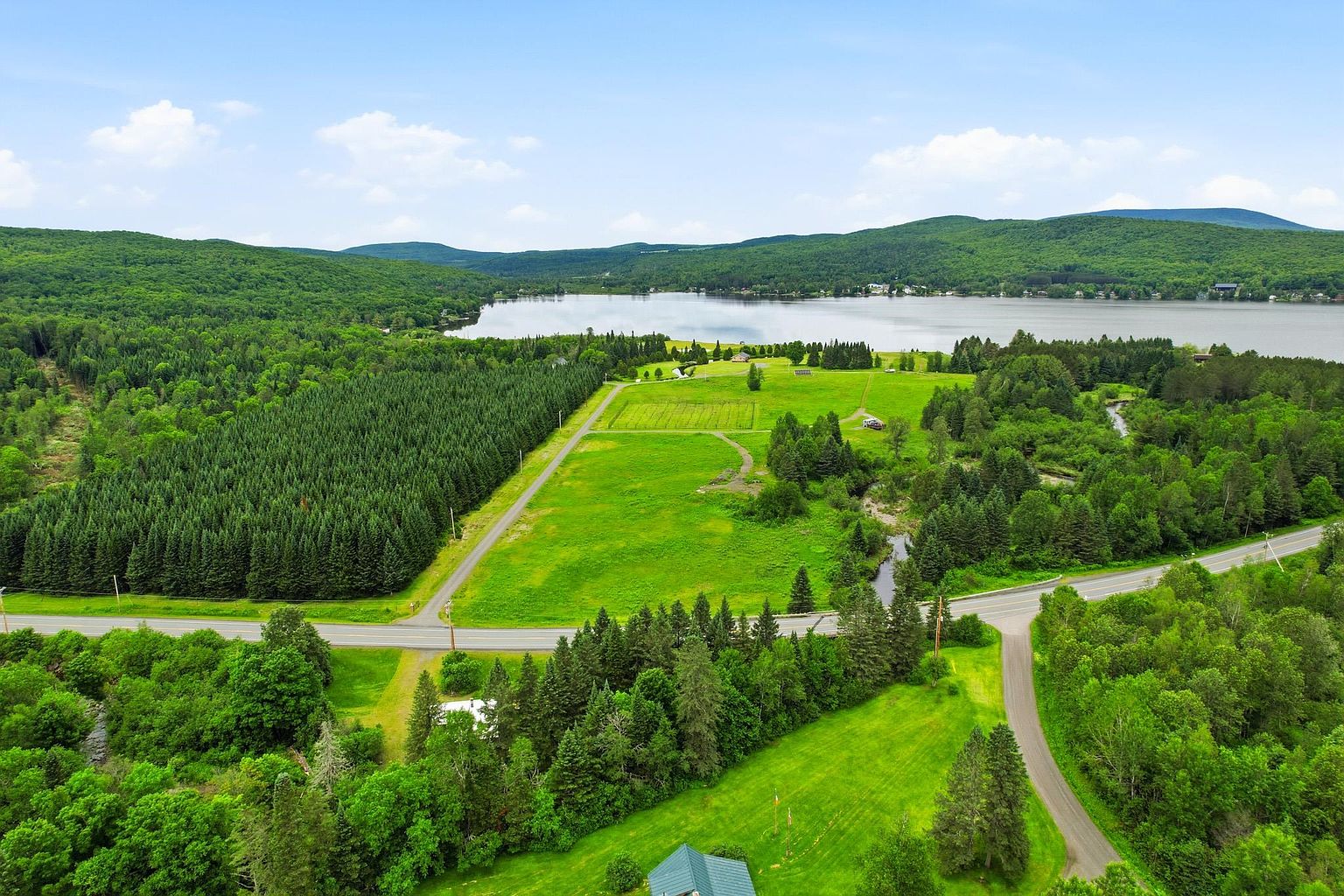 This high-angle aerial view captures a sprawling rural landscape featuring a large, open green field bordered by a dense forest and a paved road. In the distance, a serene lake stretches across the horizon, framed by rolling, tree-covered mountains under a bright, partly cloudy sky. The perspective highlights the expansive nature of the property and its peaceful, natural surroundings.