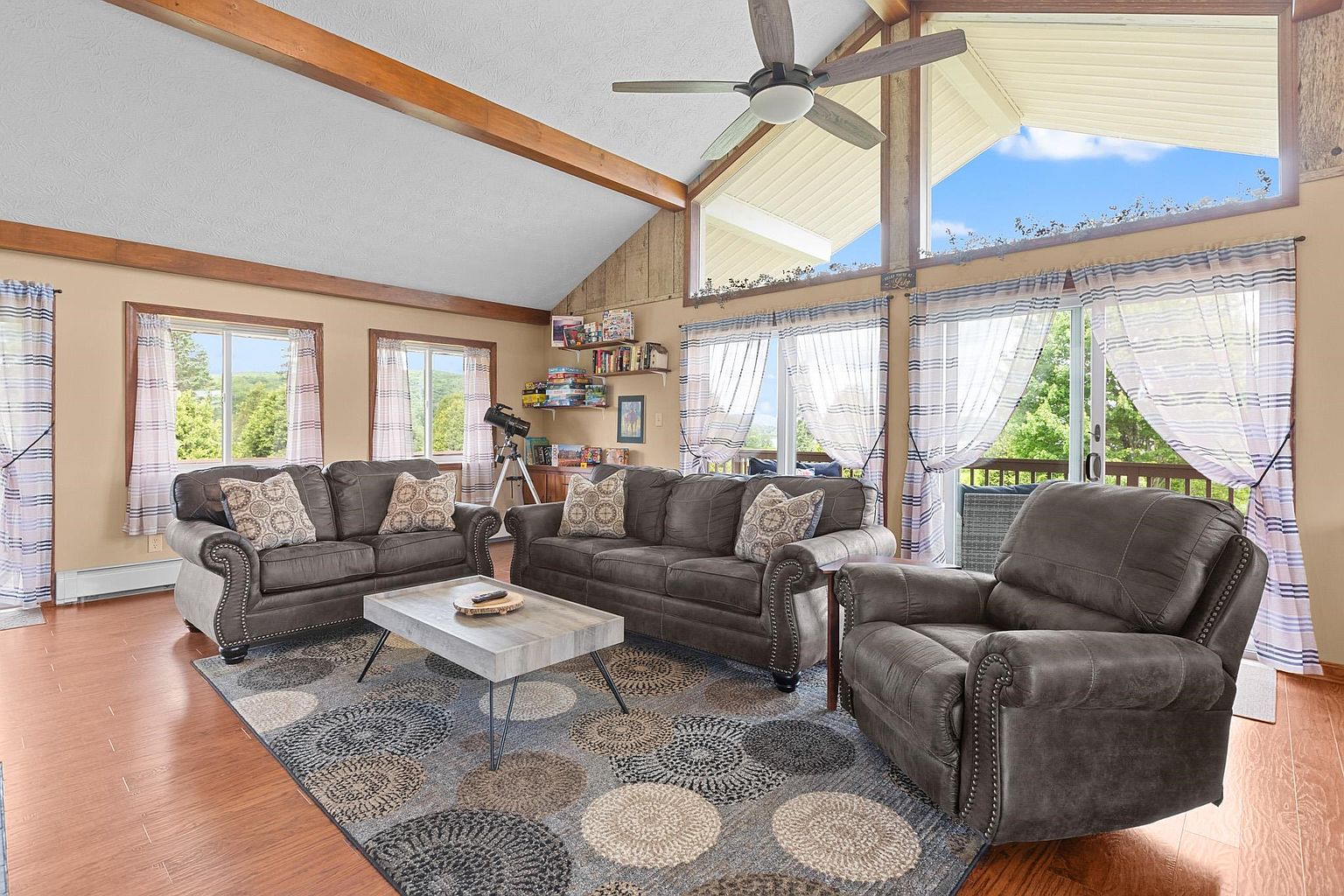This spacious living room features a vaulted ceiling with exposed wooden beams and a large ceiling fan. The room is furnished with a matching dark gray sofa set and a central coffee table, all arranged on a large patterned area rug. Large windows and glass sliding doors provide ample natural light and views of the outdoors, creating an open and inviting atmosphere.