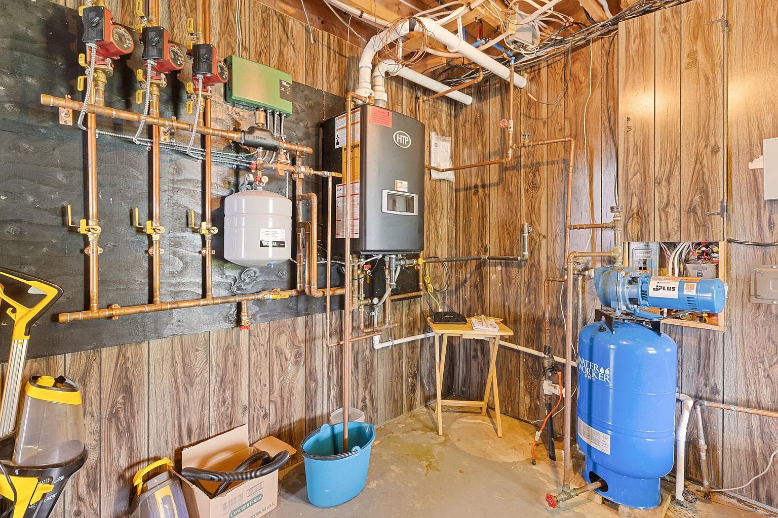 This utility area in a basement features a complex mechanical system, including an HTP wall-mounted boiler, multiple circulating pumps, and a blue Water Worker pressure tank. The equipment is mounted against wood-paneled walls, with exposed copper piping and wiring overhead. A small folding table and a blue bucket are positioned on the concrete floor, suggesting a functional, utilitarian space.