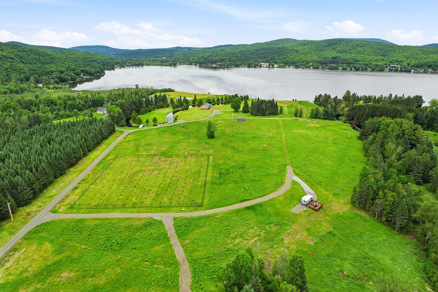 This expansive aerial view captures a large, lush green property situated along the edge of a serene lake. The landscape features a gravel driveway leading to a small recreational vehicle, a fenced-in agricultural or garden area, and a distant house nestled near the water's edge. The scene is framed by dense, mature forests and rolling hills, offering a peaceful and secluded rural atmosphere.