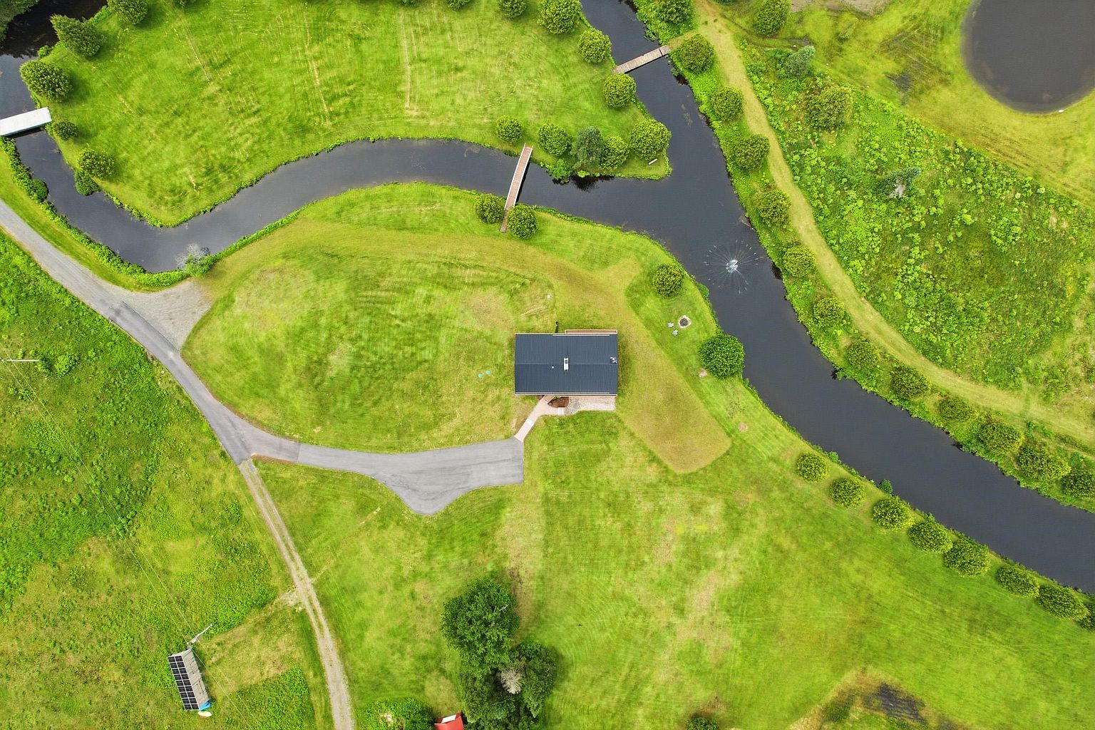 This high-angle aerial view captures a modern home situated on a sprawling, lush green property defined by a winding creek and mature landscaping. The scene features a paved driveway leading to the house, a small footbridge crossing the water, and a solar panel array in the distance. The perspective provides a comprehensive look at the expansive grounds and the serene, natural setting of the residence.