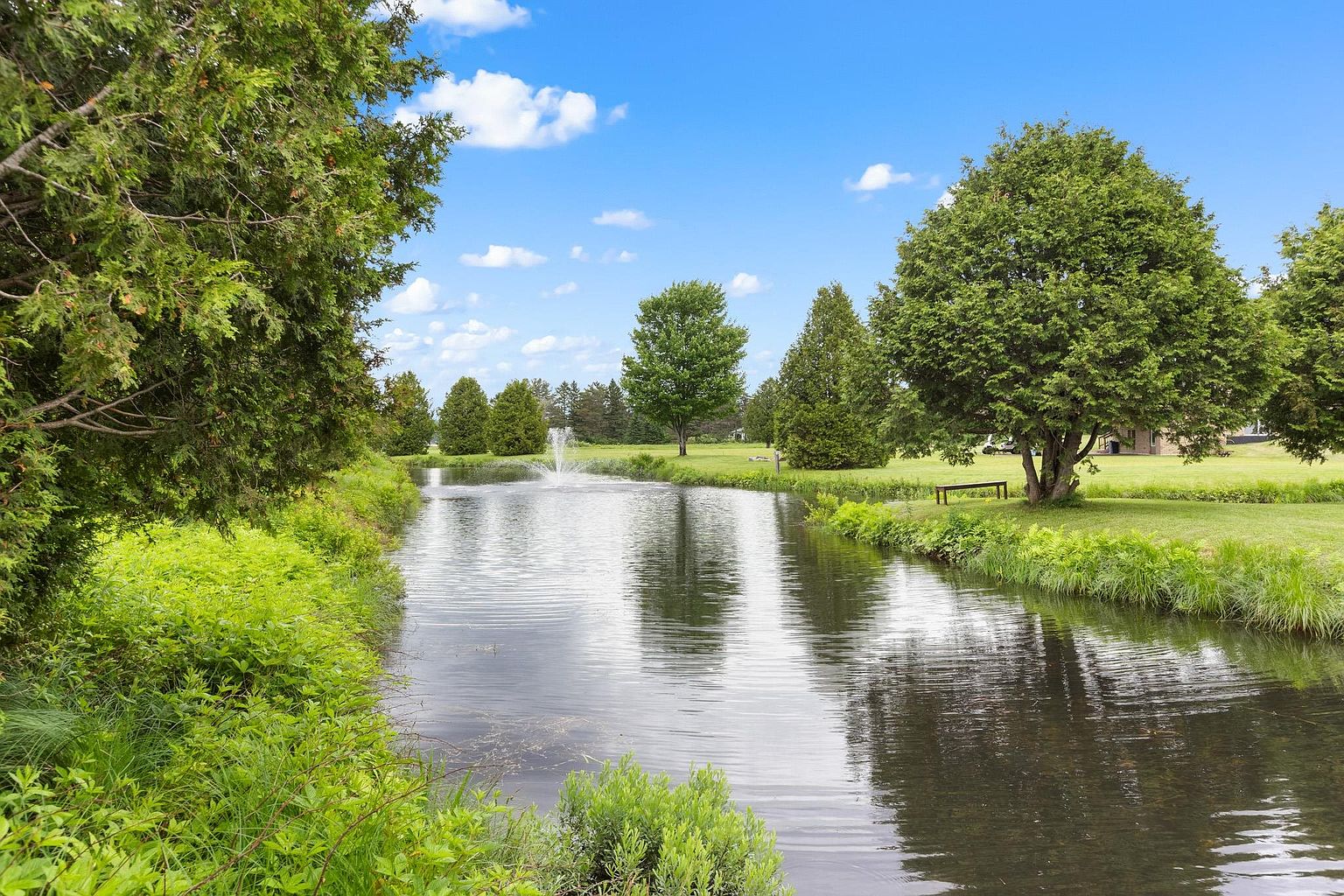 This serene outdoor scene features a tranquil pond with a central fountain, surrounded by lush green lawns and mature trees. The perspective is from the water's edge, looking across the pond toward a manicured landscape that includes a wooden bench nestled under a large tree. The bright blue sky and vibrant greenery create a peaceful and inviting atmosphere, highlighting the property's expansive and well-maintained grounds.