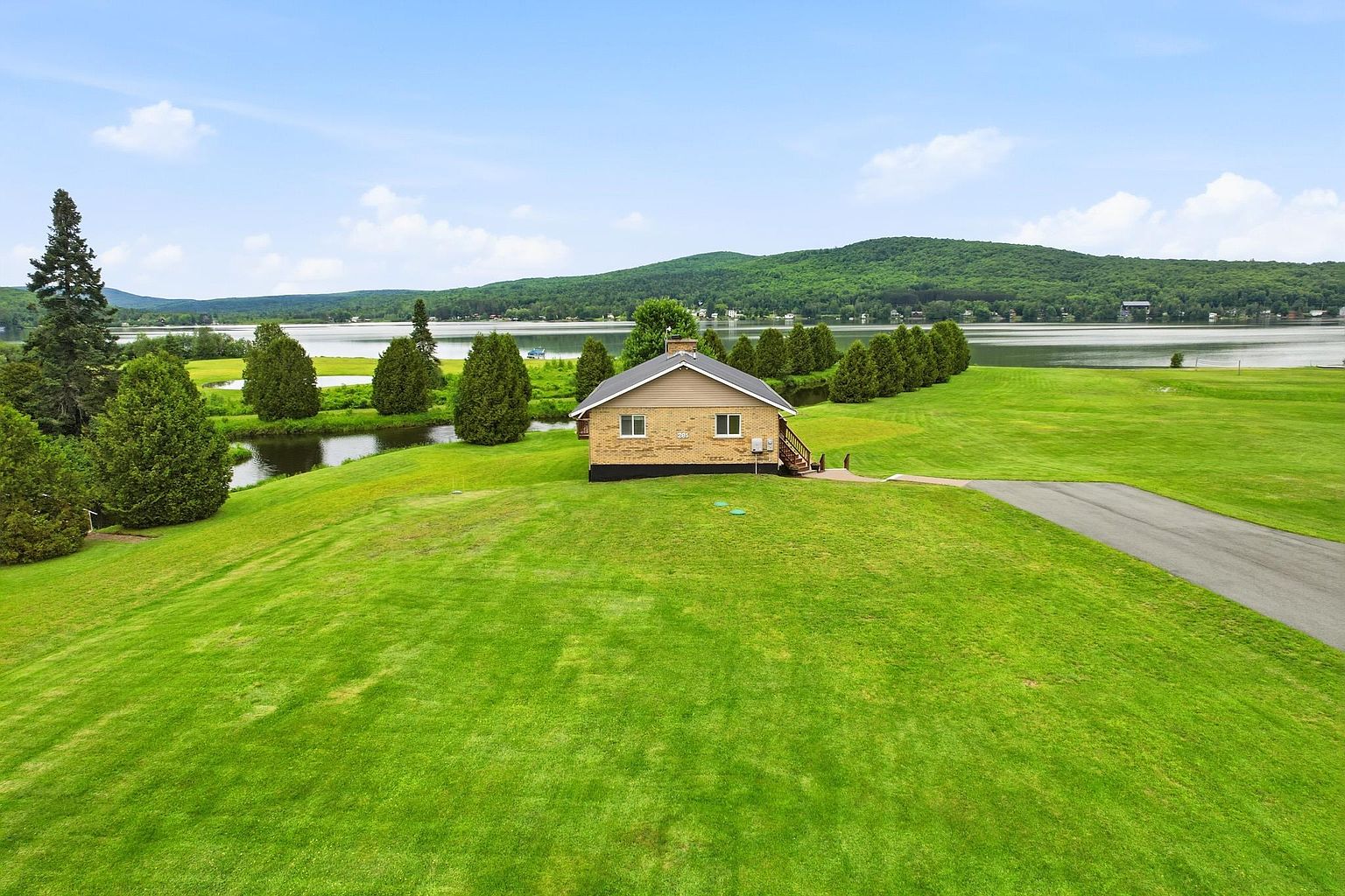 This aerial perspective captures a single-story brick home situated on a vast, manicured green lawn overlooking a serene lake. The property features a paved driveway leading to the house, which is framed by a line of mature evergreen trees and a small stream. The expansive landscape and waterfront location create a peaceful, open atmosphere ideal for outdoor living.