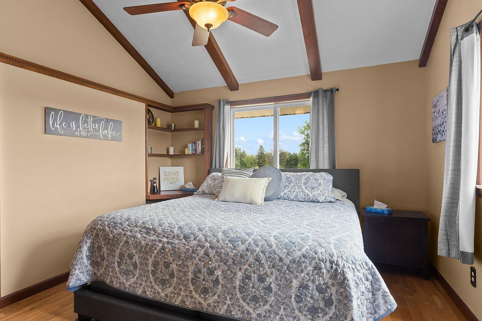 This cozy bedroom features a queen-sized bed with a blue and white patterned quilt, centered against a window that allows natural light to fill the space. To the left, a built-in wooden shelving unit provides storage and display space, complemented by a wall sign that adds a personal touch. The room is finished with warm-toned walls, exposed wooden ceiling beams, and a ceiling fan, creating a comfortable and inviting atmosphere.