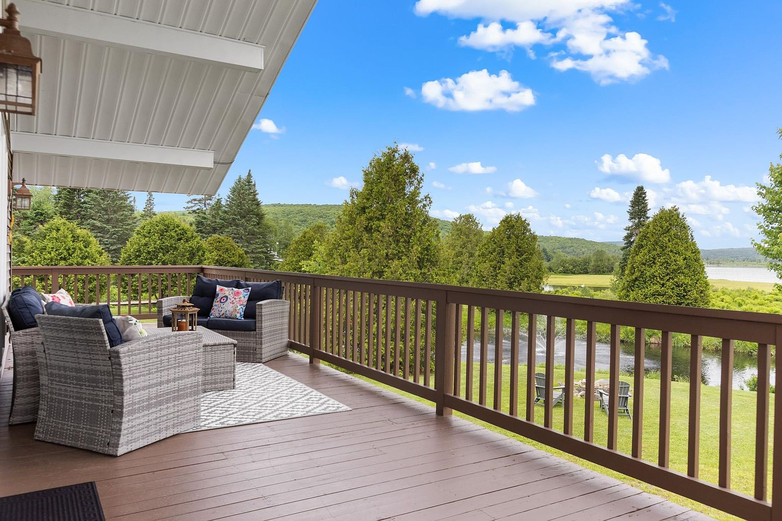 This elevated wooden deck offers a serene outdoor living space furnished with comfortable wicker-style seating and a patterned area rug. The deck features a sturdy wooden railing that overlooks a lush green lawn, a small pond with a fountain, and a scenic backdrop of rolling hills and trees under a bright blue sky. The perspective captures the inviting atmosphere of the space, highlighting its potential for relaxation and enjoying the natural landscape.