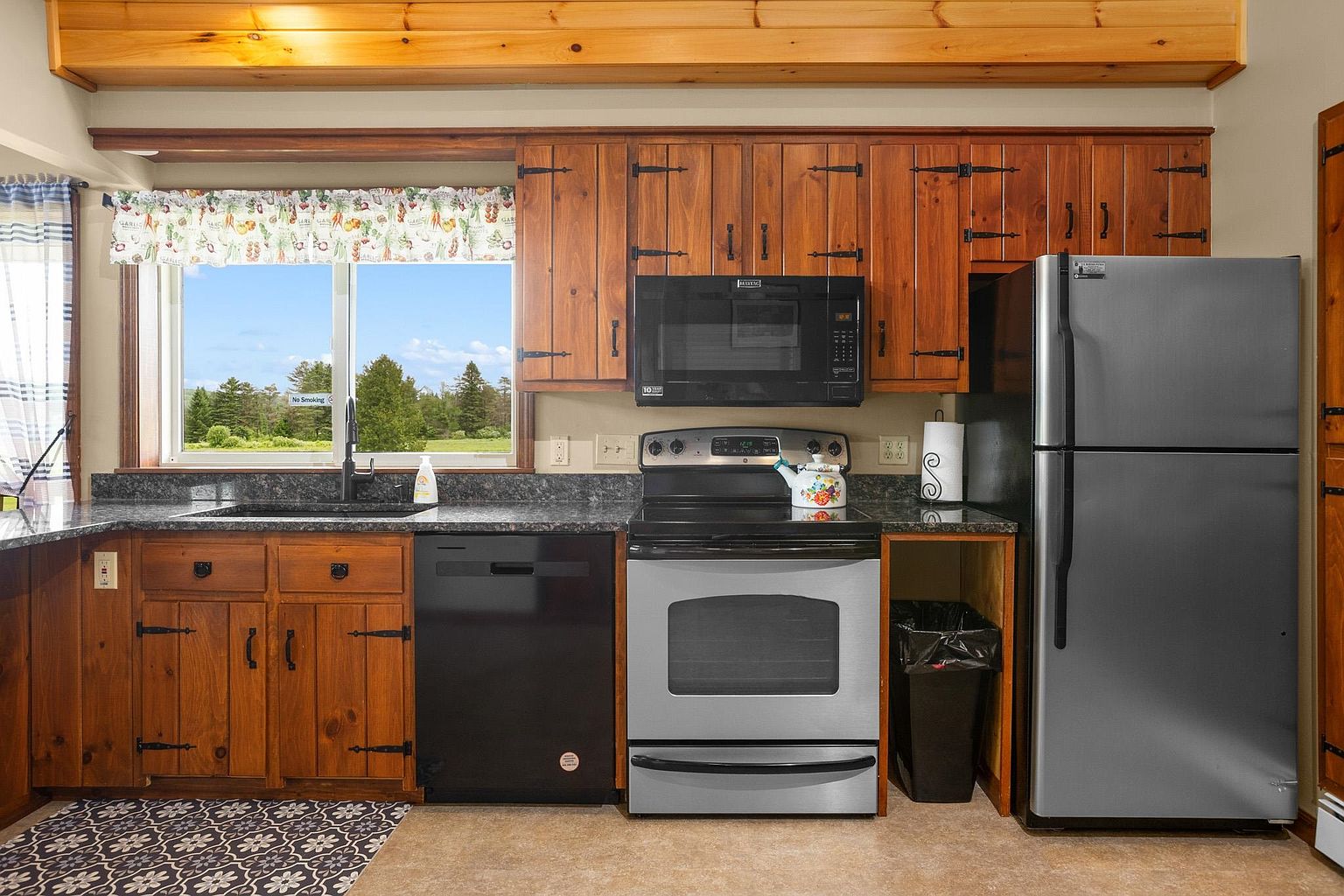 This rustic-style kitchen features warm wood cabinetry with dark hardware and a dark stone countertop. The space is equipped with stainless steel appliances, including a dishwasher, range, and refrigerator, and includes a window above the sink that provides a view of the outdoors. A patterned rug adds a decorative touch to the floor, while a wooden beam runs across the ceiling, enhancing the cabin-like aesthetic.