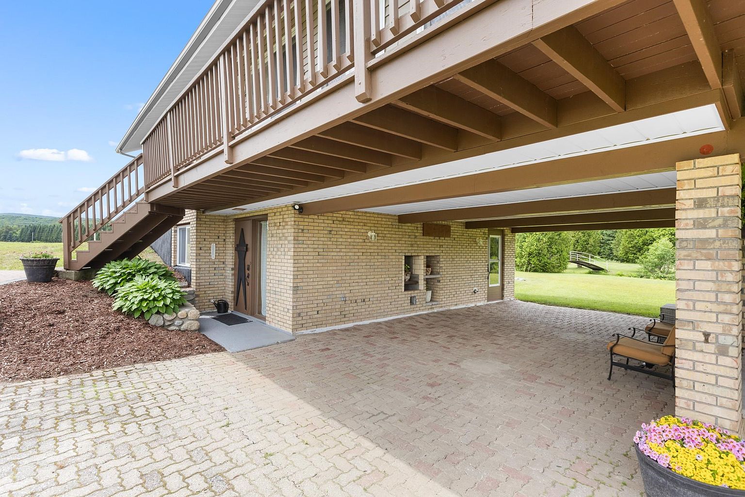 This side view of a brick home features a spacious covered patio area beneath a large wooden deck. A set of stairs leads up to the deck, while the ground level is paved with interlocking bricks, creating a functional outdoor living space. The scene is framed by a brick pillar, a potted plant with colorful flowers, and a view of the surrounding lawn and landscape.