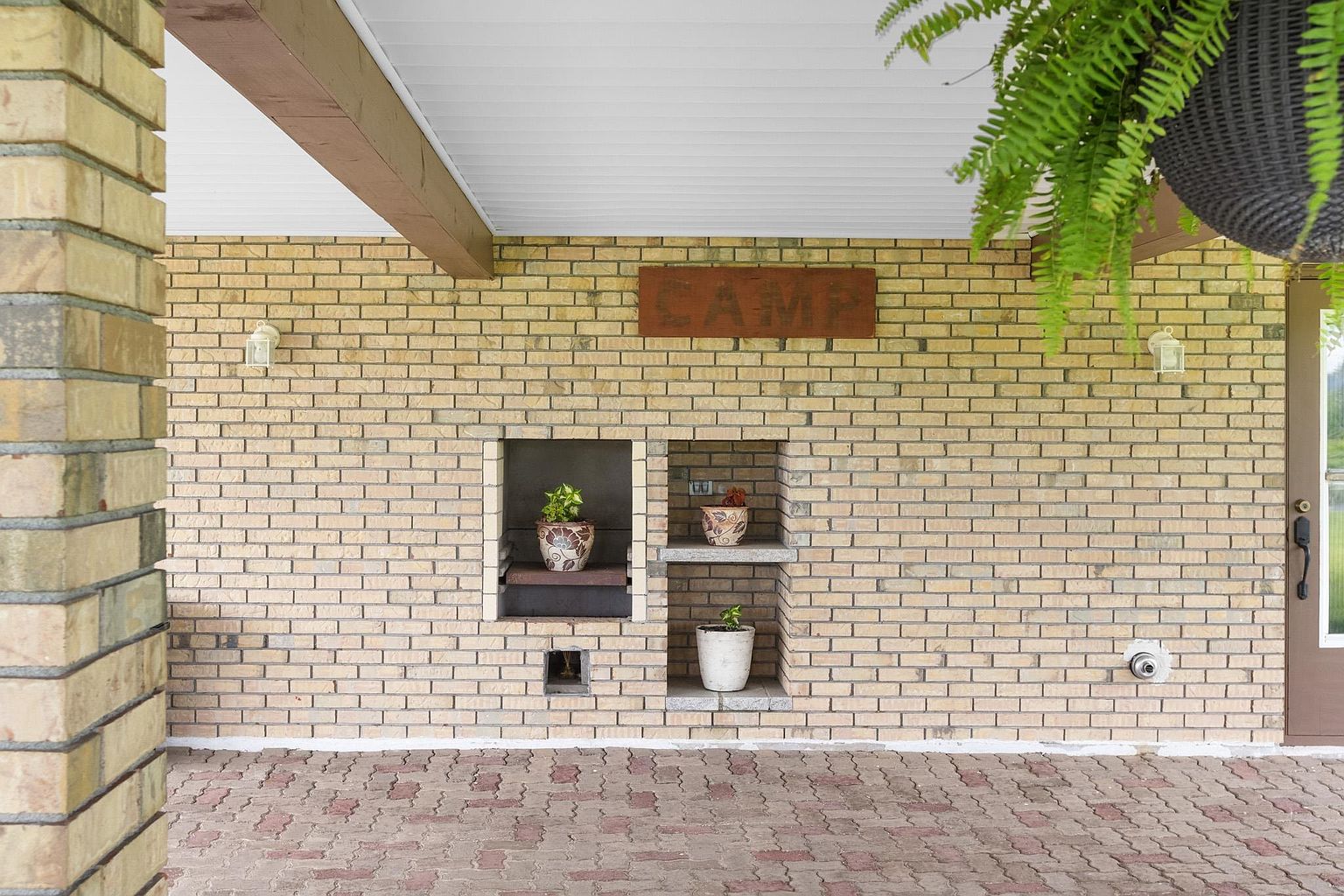 This covered patio area features a light-colored brick wall adorned with a rustic wooden 'CAMP' sign and two recessed niches used for decorative potted plants. The ground is paved with reddish-brown brick pavers, and a hanging fern adds a touch of greenery to the space. The perspective is a straight-on shot, highlighting the textured brickwork and the inviting, sheltered outdoor living area.
