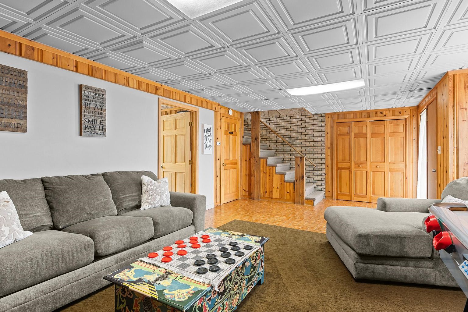 This finished basement features a cozy seating area with two grey sofas arranged around a colorful, custom-painted coffee table designed as a checkerboard. The space includes wood-paneled walls, a drop-tile ceiling, and a staircase leading to the upper level, which is accented by a brick wall. The room offers a casual, multi-functional layout suitable for recreation and relaxation.