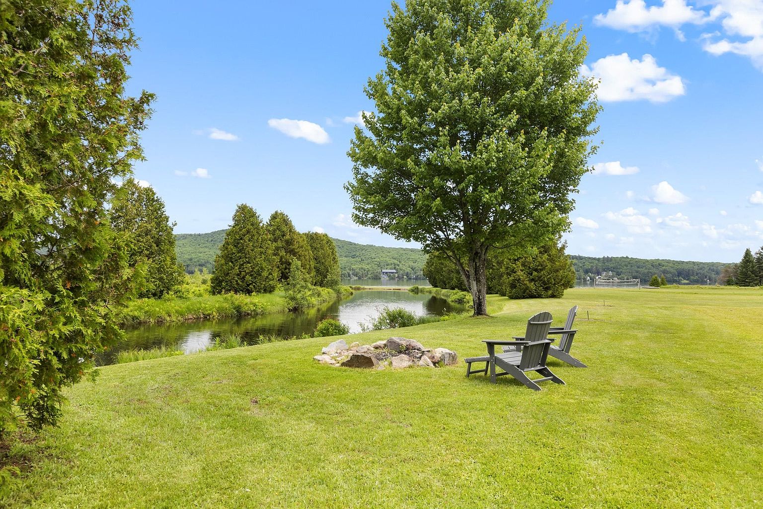 This serene outdoor space features a lush, expansive green lawn leading down to a calm, reflective pond bordered by mature trees. A stone fire pit sits prominently in the foreground, accompanied by two Adirondack chairs that offer a peaceful spot to enjoy the waterfront view. The scene is captured from a slightly elevated perspective, showcasing the natural beauty of the landscape under a bright, partly cloudy sky.