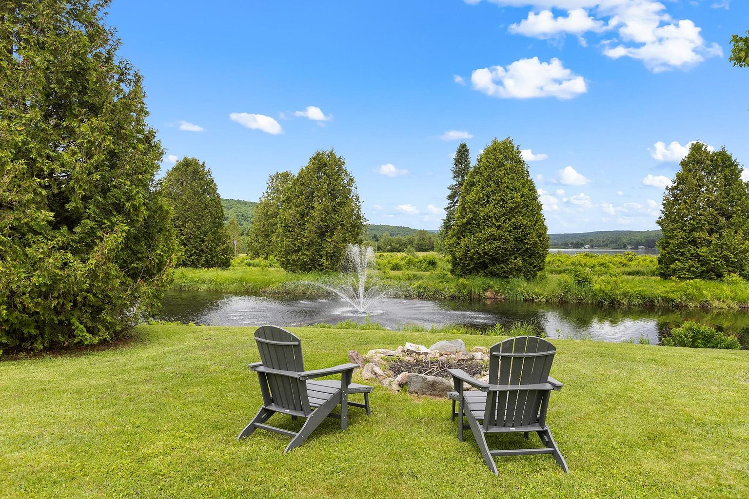 This serene backyard scene features a lush green lawn with two dark Adirondack chairs positioned around a stone-lined fire pit. In the background, a decorative water fountain sprays into a tranquil pond, framed by mature evergreen trees and a scenic landscape. The perspective is a wide, eye-level shot that captures the peaceful atmosphere and natural beauty of the property's outdoor space.