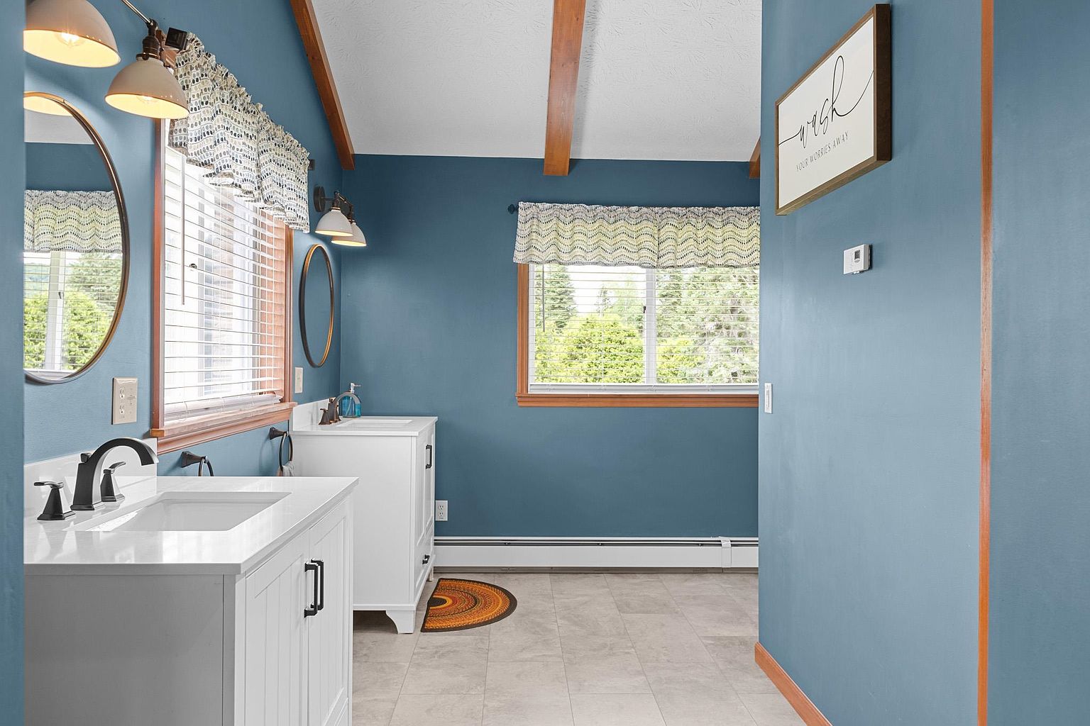 This bright bathroom features a dual-vanity setup with white cabinetry and dark hardware against bold blue walls. The space is accented by warm wood-toned trim and exposed ceiling beams, while patterned valances over the windows add a touch of character. A framed sign on the wall and light-colored tile flooring complete the inviting, well-lit aesthetic.