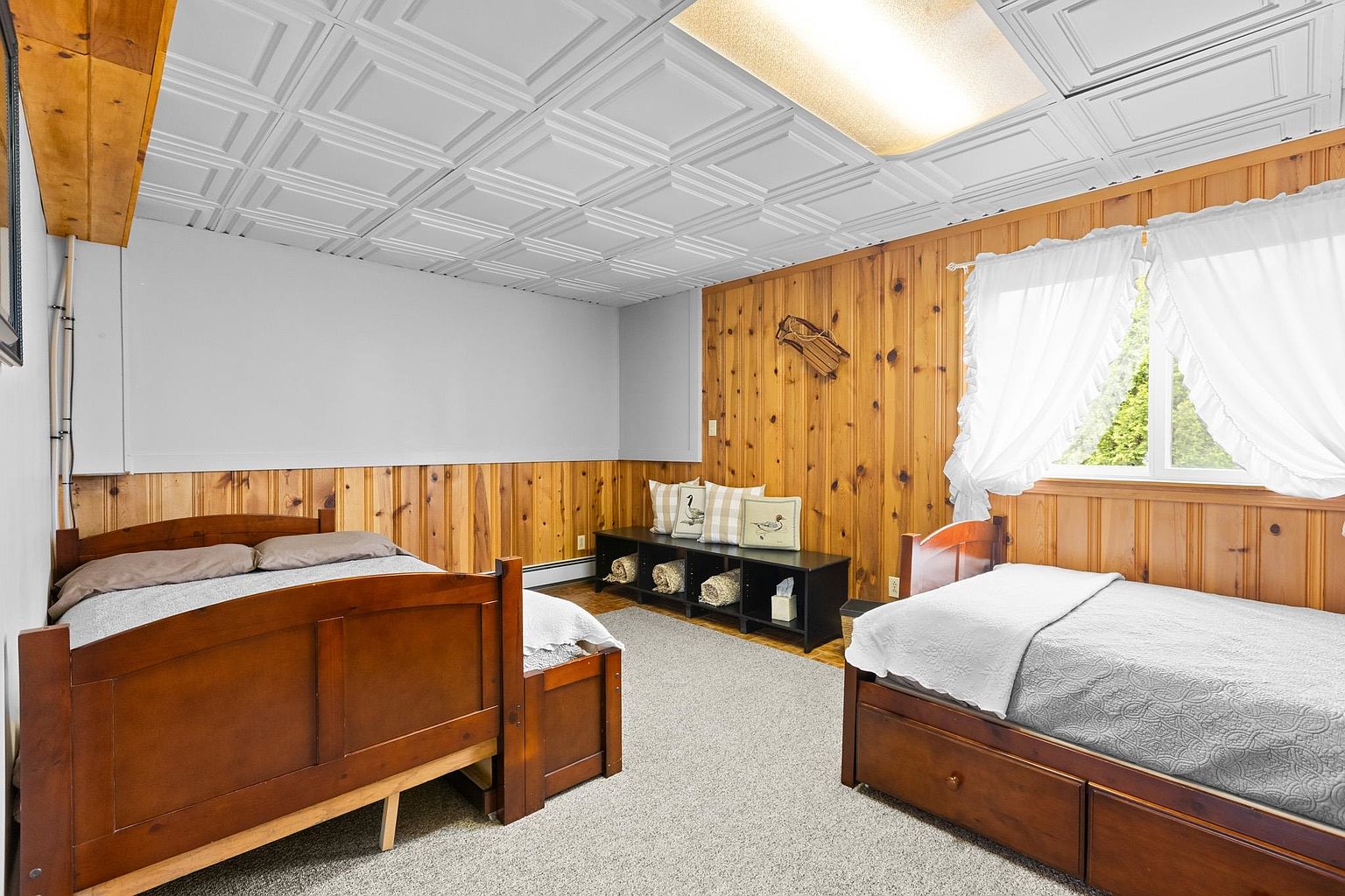 This bedroom features two wooden bed frames set against a combination of light-colored walls and warm wood-paneled wainscoting. A low black storage unit with baskets sits between the beds, topped with decorative pillows, while a window with white curtains allows natural light into the space. The room is finished with a textured ceiling and neutral carpeting, creating a cozy and rustic atmosphere.