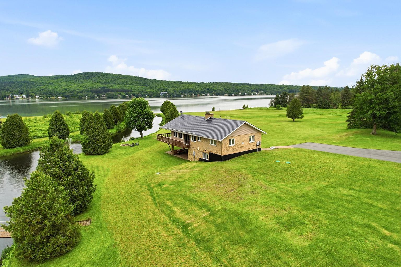 This aerial perspective captures a brick-clad home situated on a sprawling, lush green lawn overlooking a serene lake. The property features a dark metal roof, a wooden deck extending from the rear, and a paved driveway leading toward the structure. The surrounding landscape is characterized by mature trees, a small winding stream, and expansive open fields, creating a tranquil and picturesque waterfront setting.