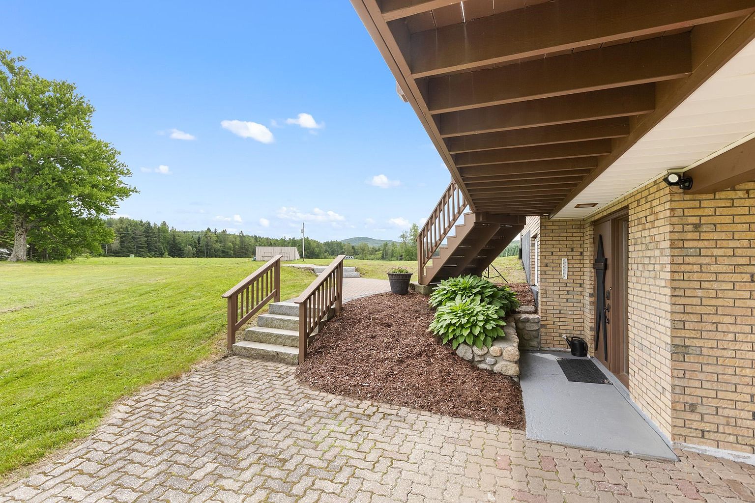This rear exterior view showcases a brick-clad home with a wooden staircase leading up to an elevated deck. A paved patio area connects to a set of stone steps that transition into a expansive, well-maintained grassy lawn. The scene is framed by a clear blue sky and a distant treeline, creating a peaceful and open outdoor living space.