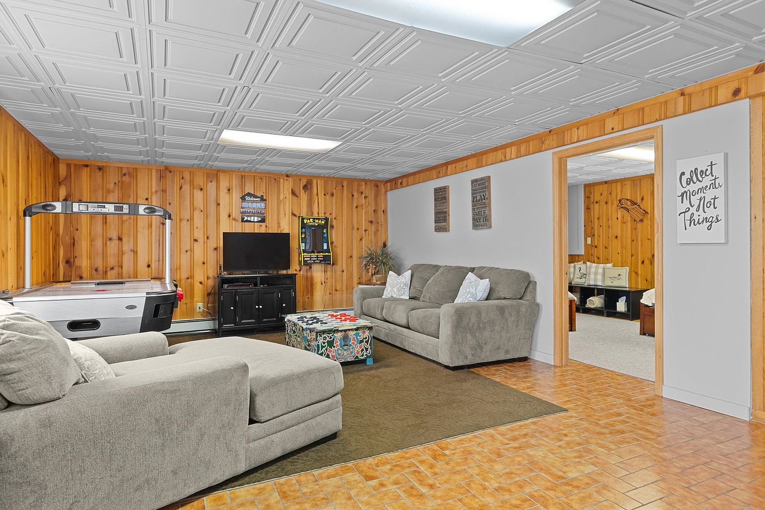 This spacious basement recreation area features wood-paneled walls, a drop ceiling, and tiled flooring, creating a cozy and casual atmosphere. The room is furnished with a large grey sectional sofa, a patterned ottoman, and an air hockey table, making it an ideal space for entertainment. A doorway leads to an adjacent room, while a wall sign adds a decorative touch to the inviting, multi-functional living space.