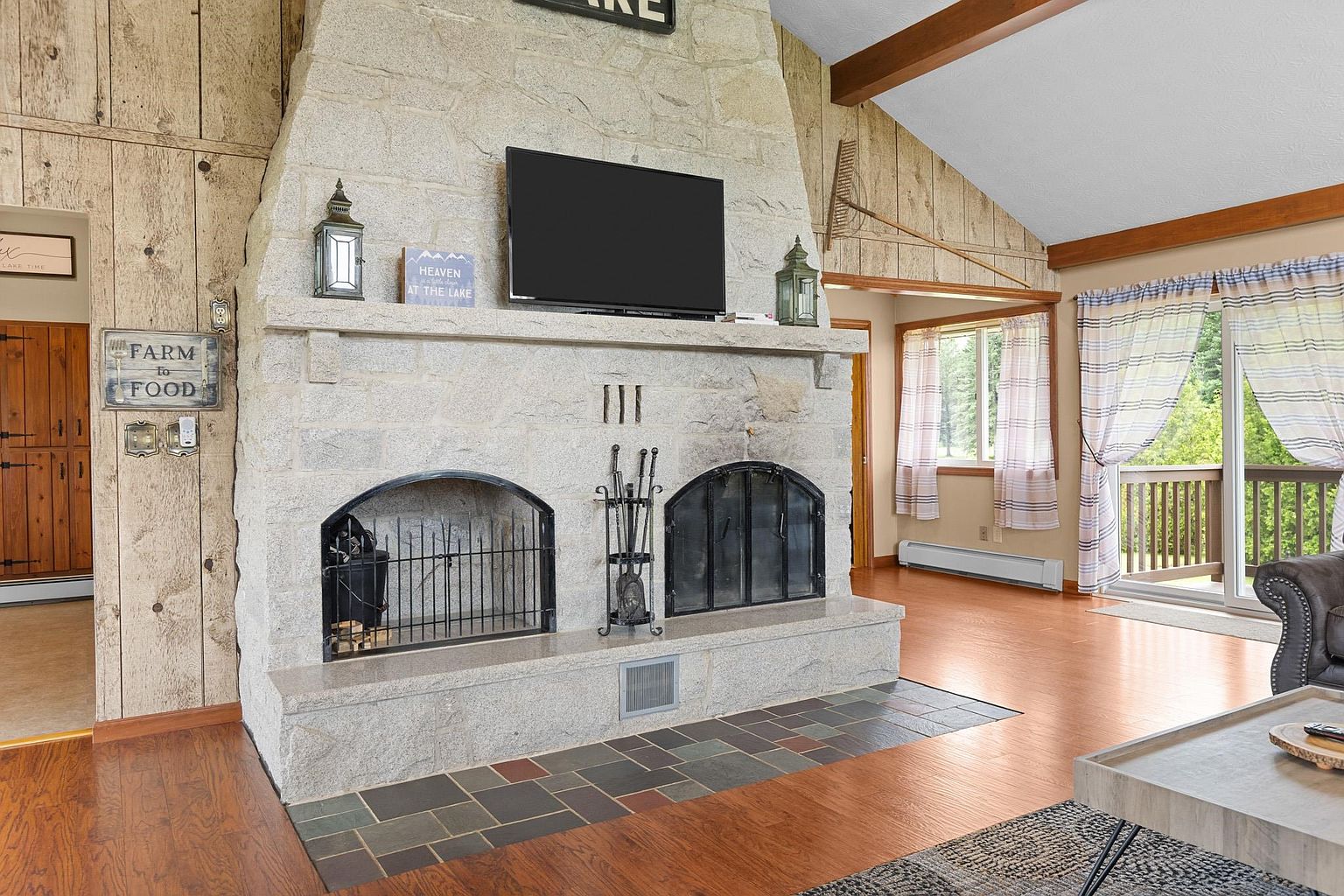 This living room features a prominent stone-clad chimney breast that serves as the focal point, housing a television and two arched fireboxes. The space is accented by rustic wood-paneled walls, a multi-colored slate hearth, and a sliding glass door that provides access to an outdoor deck. The room maintains a cozy, cabin-inspired aesthetic with warm wood flooring and exposed structural beams.