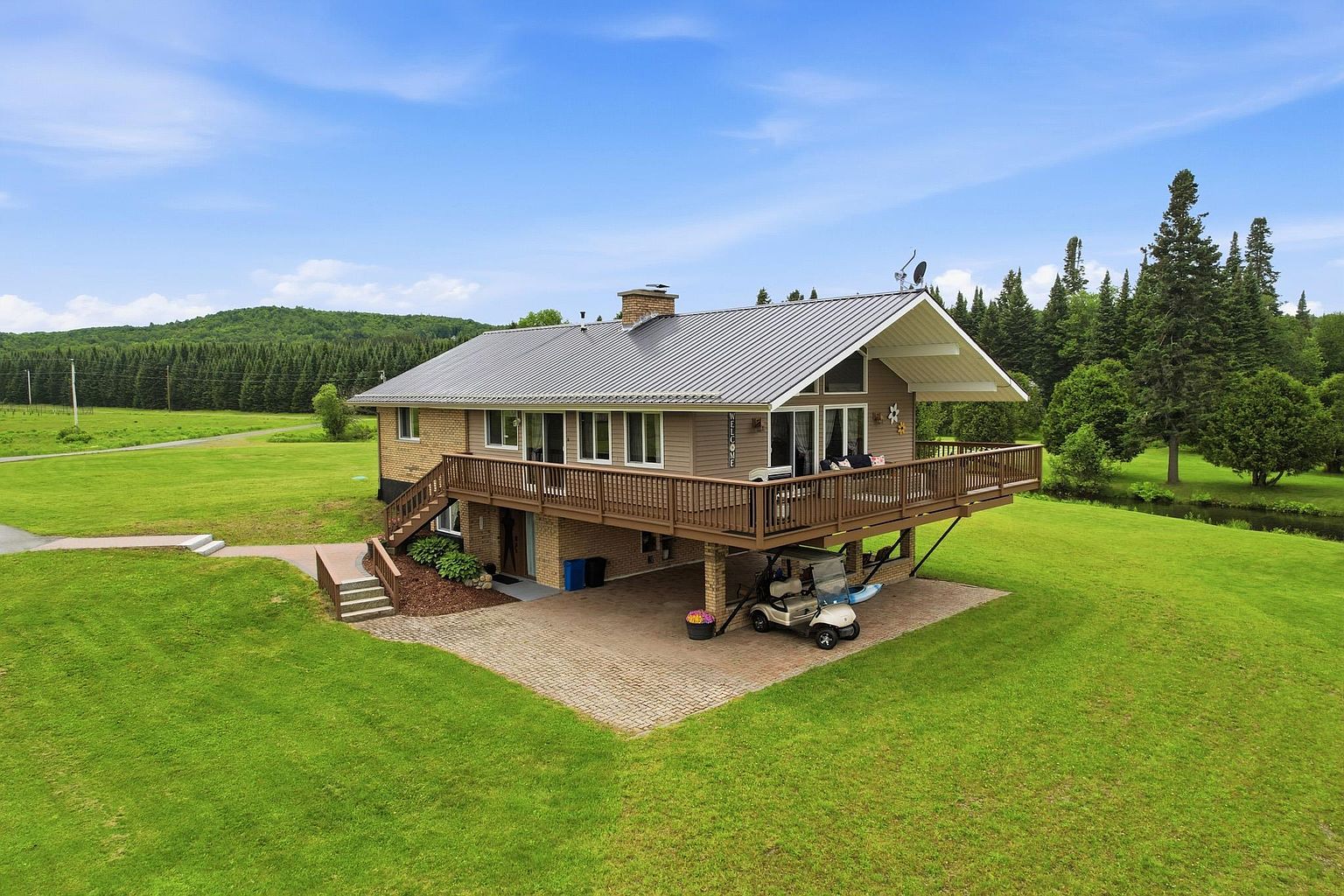 This elevated aerial perspective captures a split-level home set against a backdrop of lush green forests and rolling hills. The property features a prominent wrap-around wooden deck, a paved lower-level carport area housing a golf cart, and a brick and siding exterior. The expansive lawn and serene natural surroundings create a peaceful, rural atmosphere ideal for outdoor living.