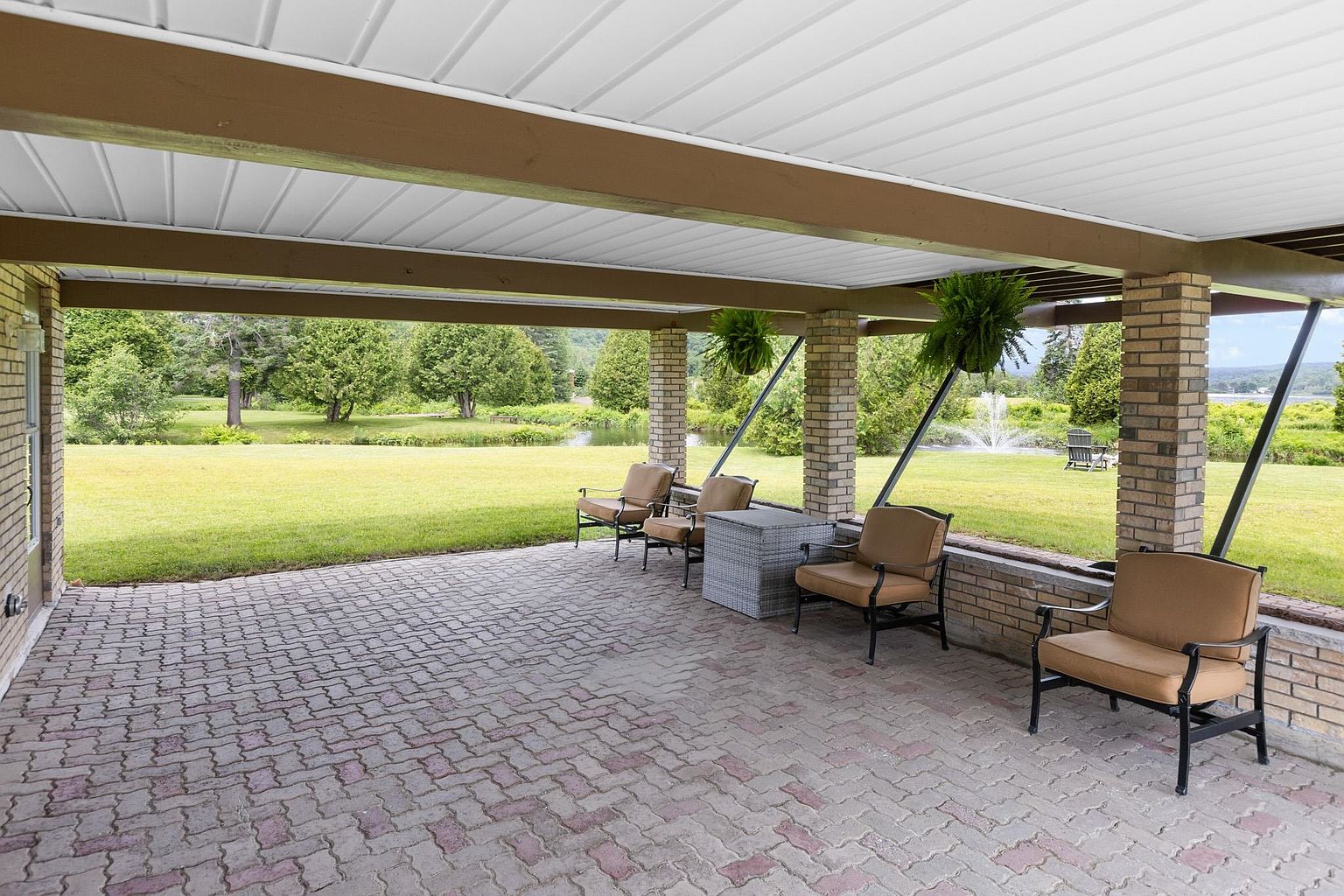 This covered patio features a classic brick-paved floor and a sturdy structure supported by brick pillars, offering a sheltered outdoor living space. The area is furnished with comfortable seating arranged around a central table, creating an inviting spot to enjoy the expansive green lawn and distant water views. Hanging ferns add a touch of natural greenery to the space, enhancing the peaceful and scenic atmosphere.