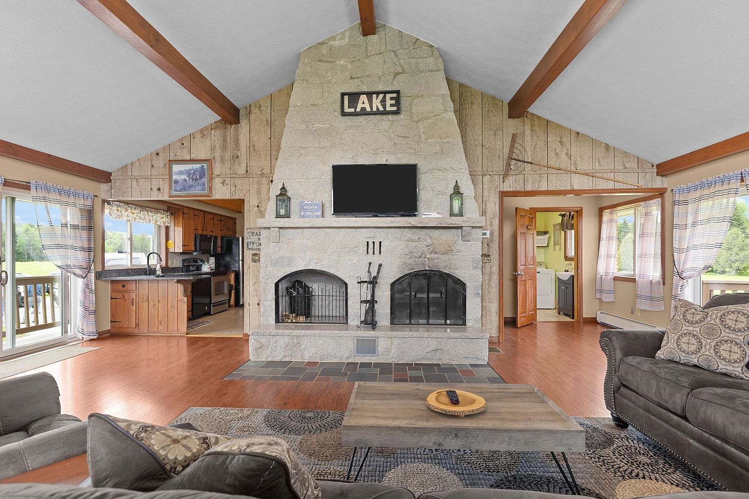 This spacious living room features a vaulted ceiling with exposed wooden beams and a prominent stone-clad chimney breast that serves as the room's focal point. The open-concept layout connects to a kitchen on the left and a laundry area on the right, while large sliding glass doors provide access to an outdoor deck. The room is furnished with comfortable seating and a rustic coffee table, creating a cozy, cabin-like atmosphere.