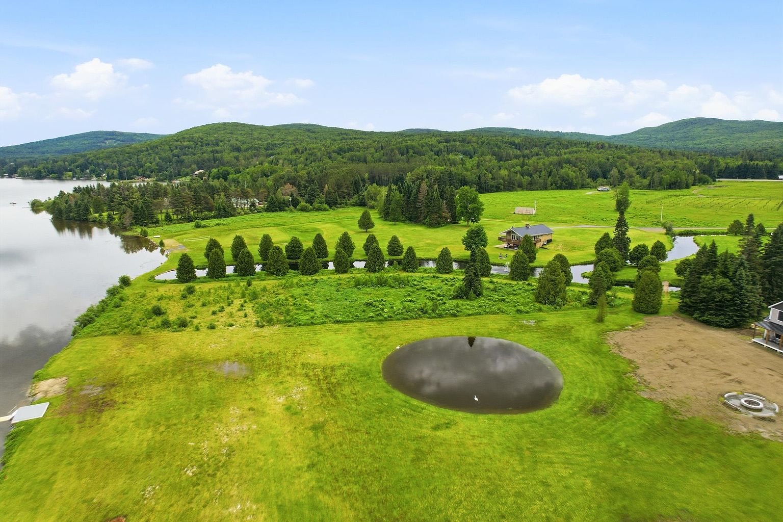 This expansive aerial view captures a sprawling rural property featuring a large, manicured green lawn, a private pond, and a line of mature evergreen trees. In the distance, a rustic cabin-style home sits nestled near a serene lake, surrounded by dense forest and rolling hills under a bright, partly cloudy sky. The perspective provides a comprehensive look at the property's vast landscape and its peaceful, secluded setting.